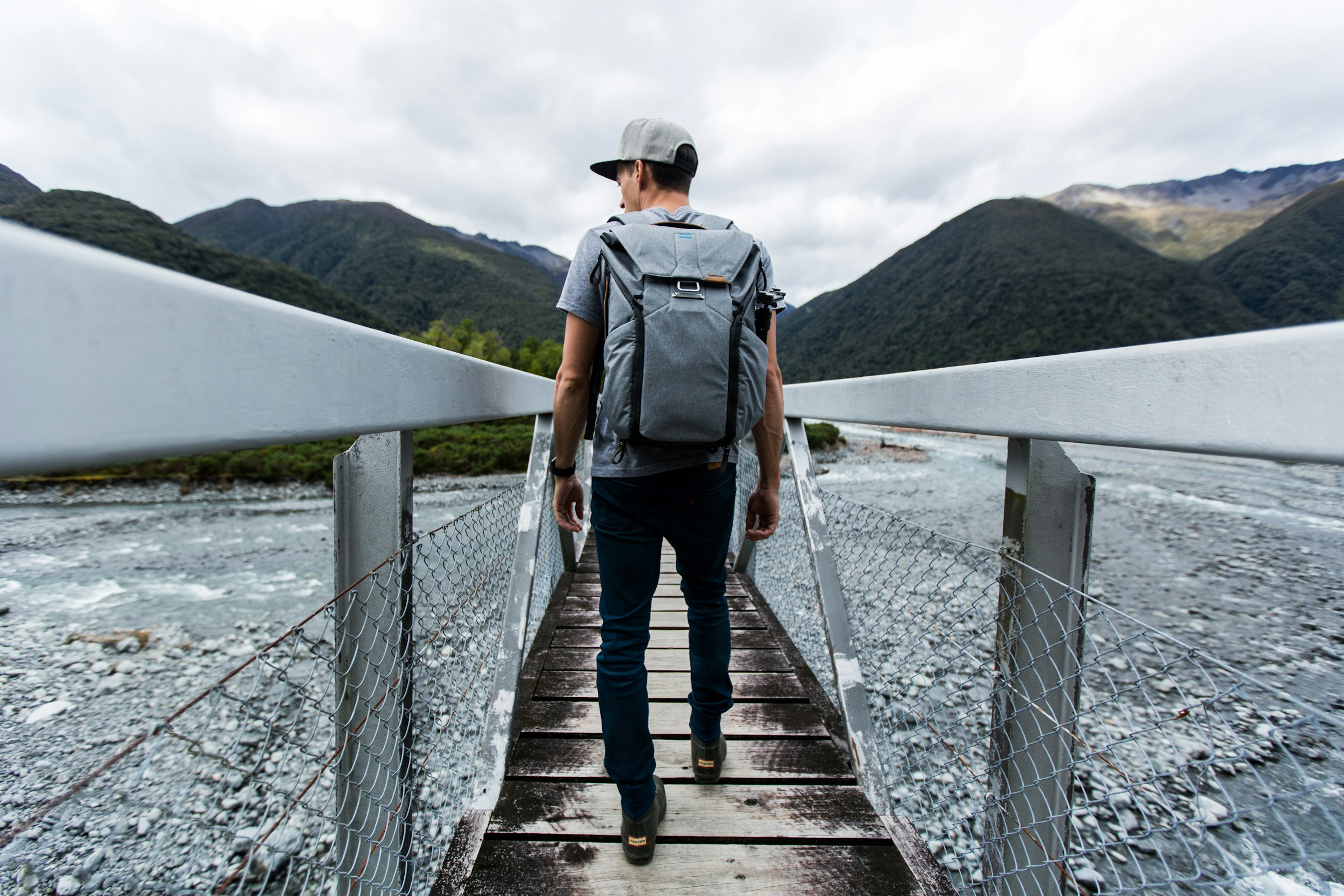 A man wearing an Everyday backpack is going across a walkway with wooden rails. The ground either side of him is covered in gravel, mountains are in the near distance.