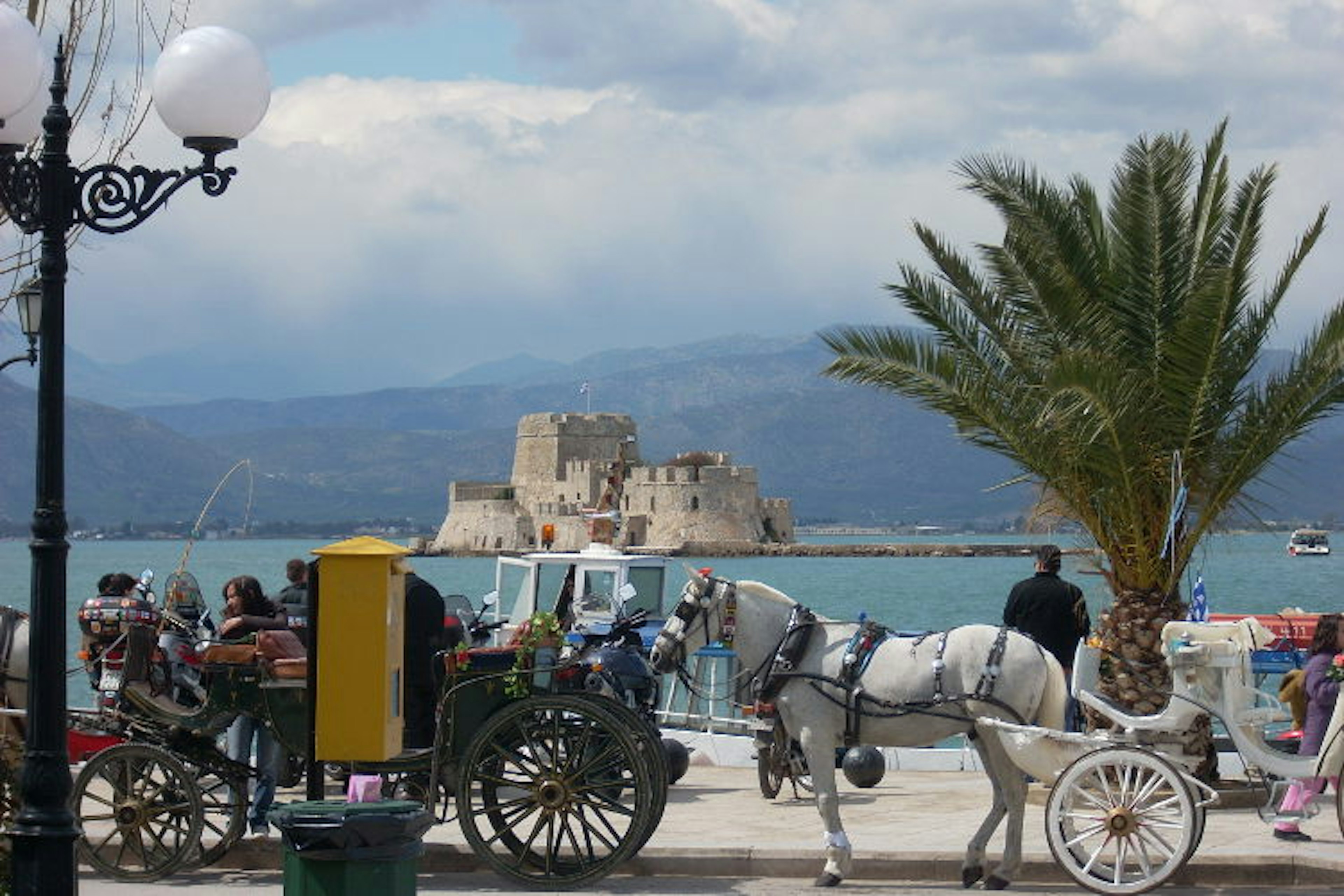 Two horse-drawn carriages stand dockside. An island fort can be seen in the bay, with large hills rising behind.