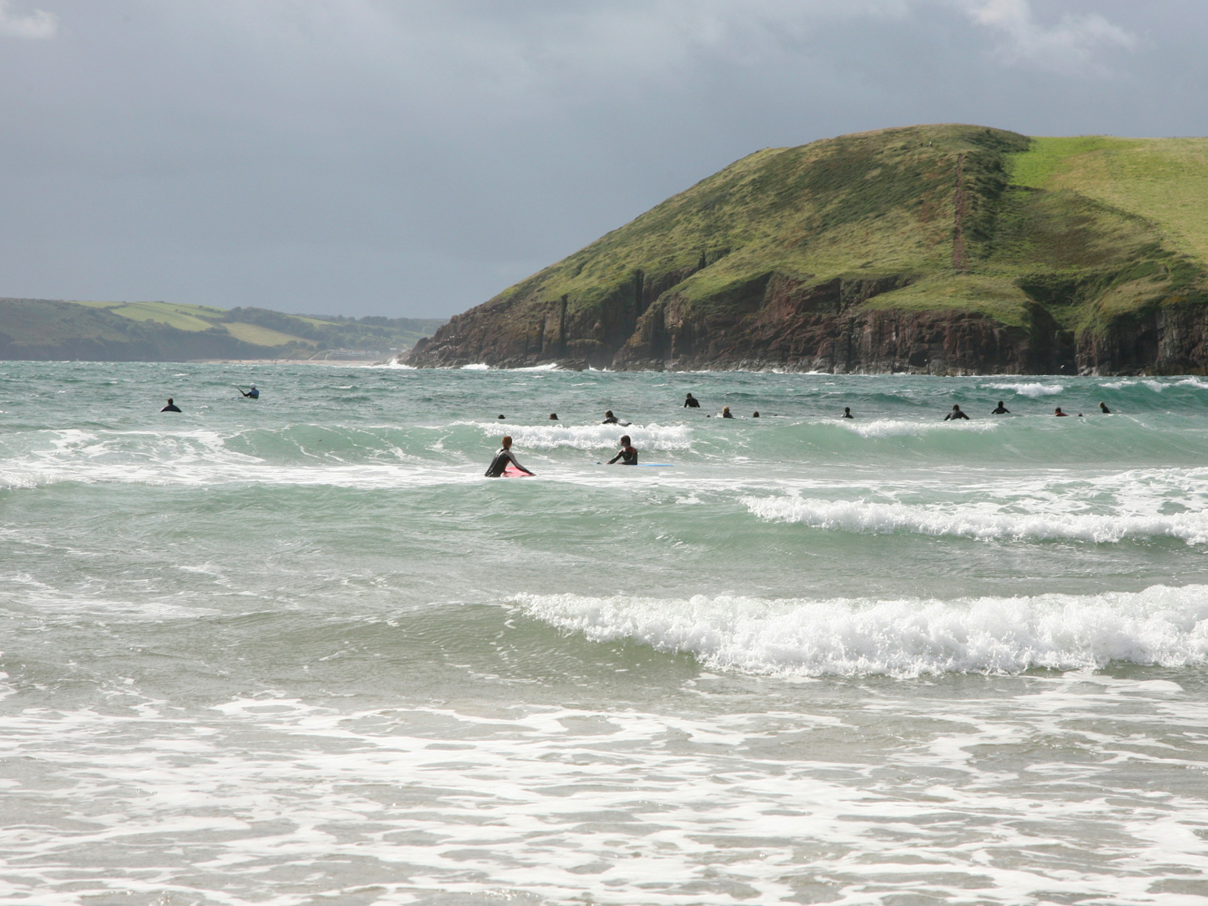 Many surfers wait in the seas for a wave, with hills of the Pembrokeshire coast green in the distance