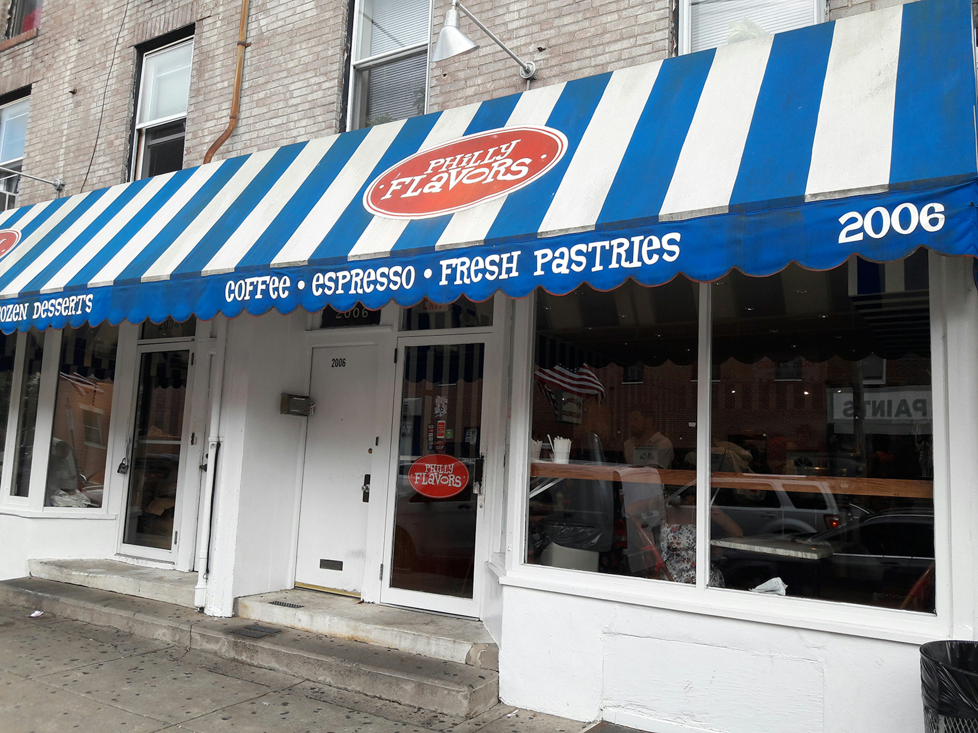 Storefront with glass windows, white trim and a blue-and-white awning that has the words ' Philly Flavors' in a red circle