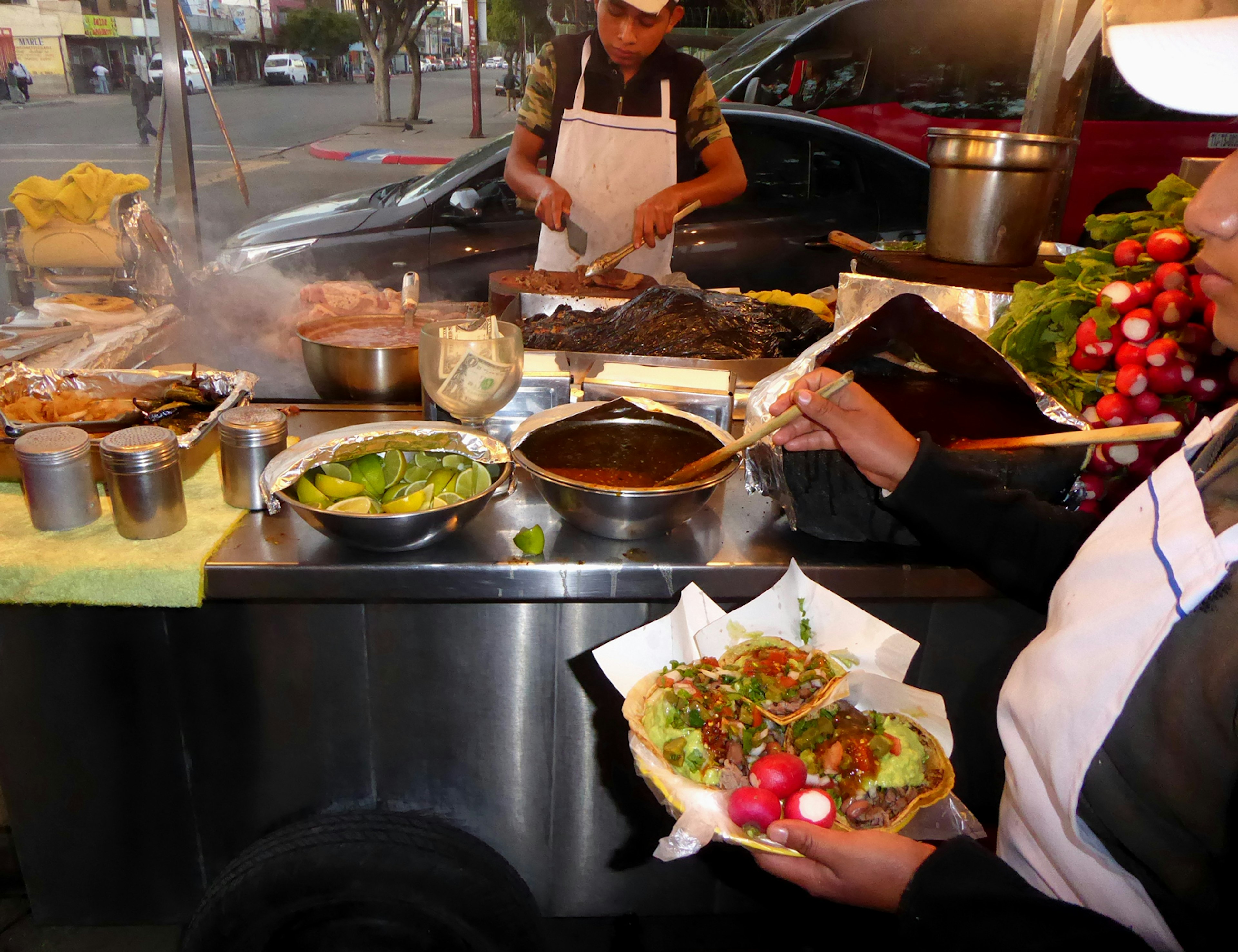 A man cooks on in a roadside stand while a woman holds a plate of tacos