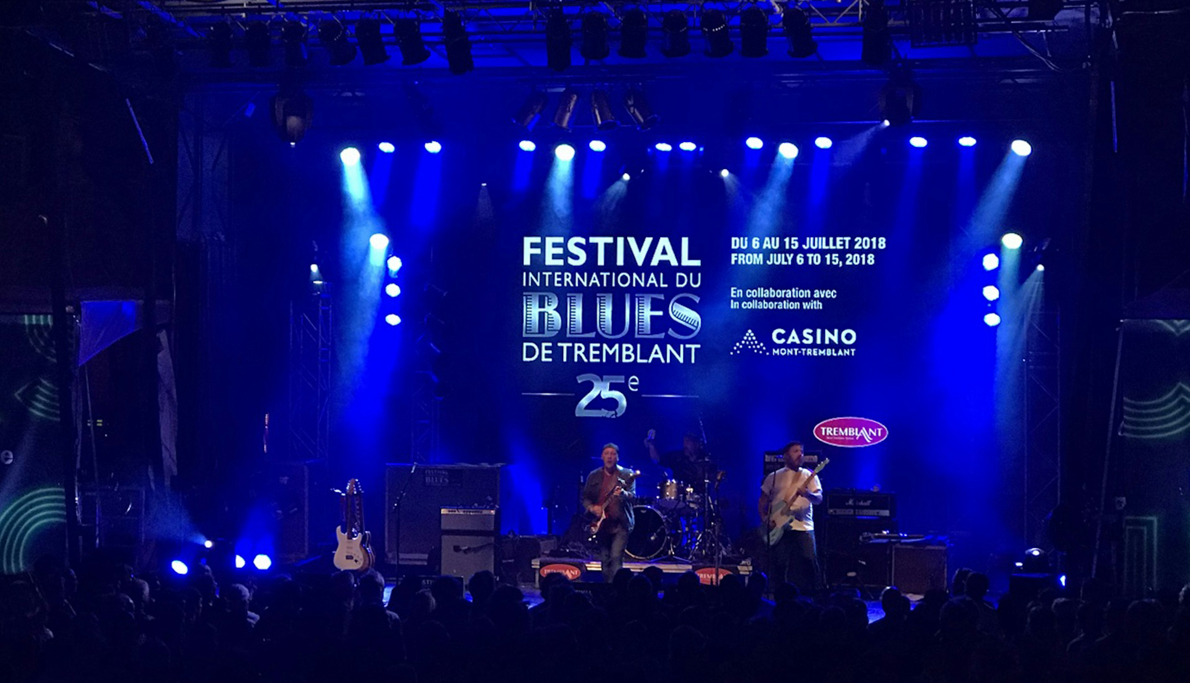 A band plays on an outdoor stage as blue lights illuminate a screen behind them that announces this is the Festival of Blues in Mont-Tremblant, Quebec.