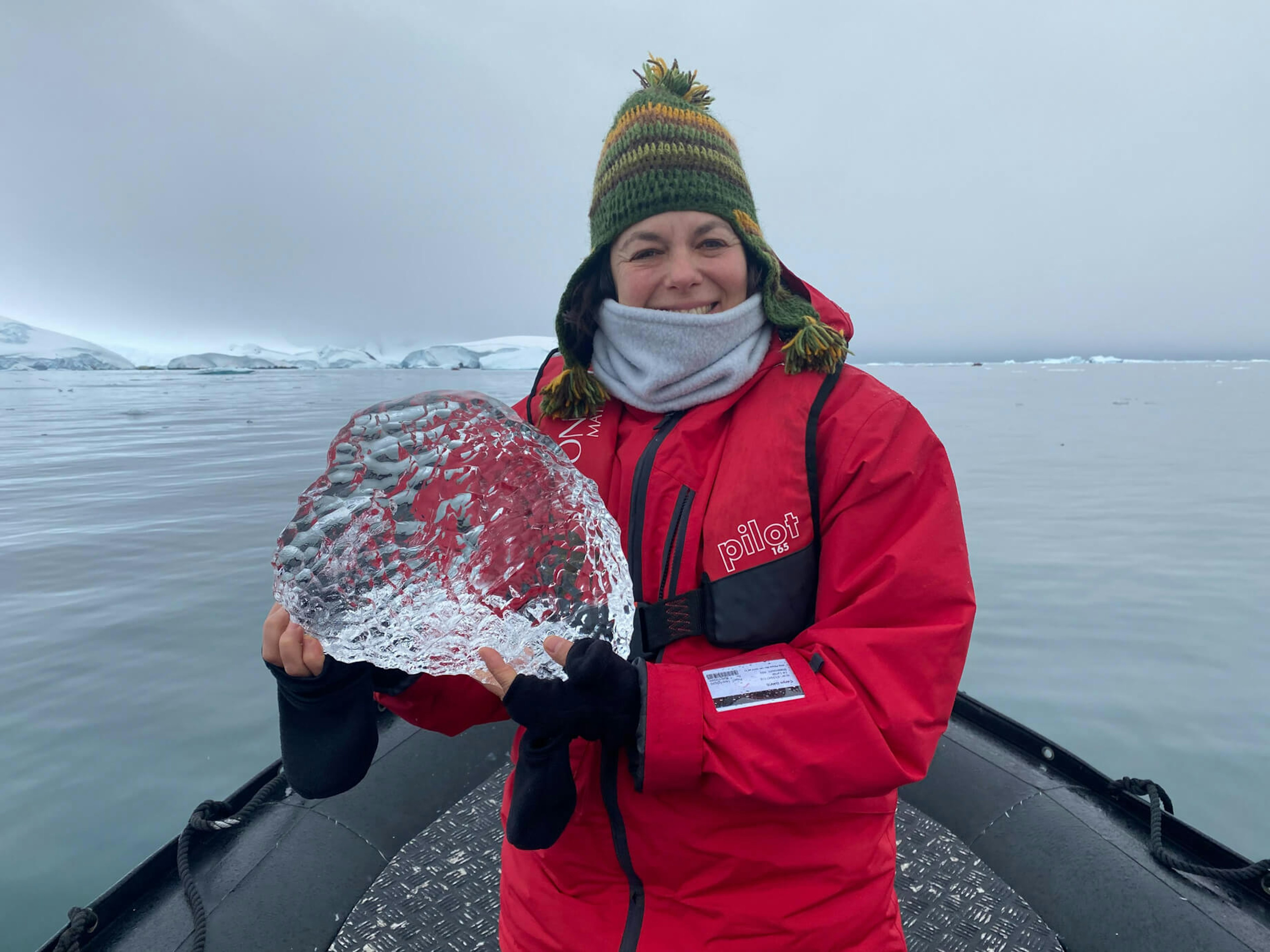 The author stands at the top of a boat, holding a sparkling piece of ice in her hands. She is wearing a red jacket, woolly hat and gloves.