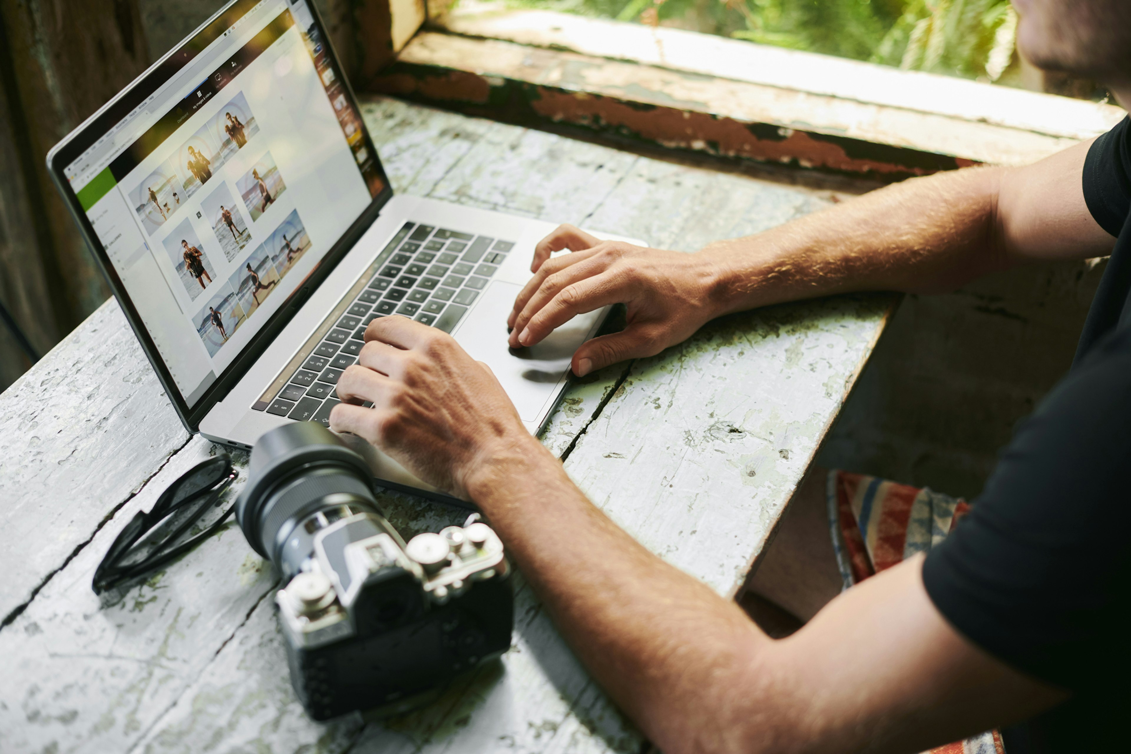 A man is sitting a worn-out table editing photos on a laptop. His camera is sitting next to the laptop on the desk.