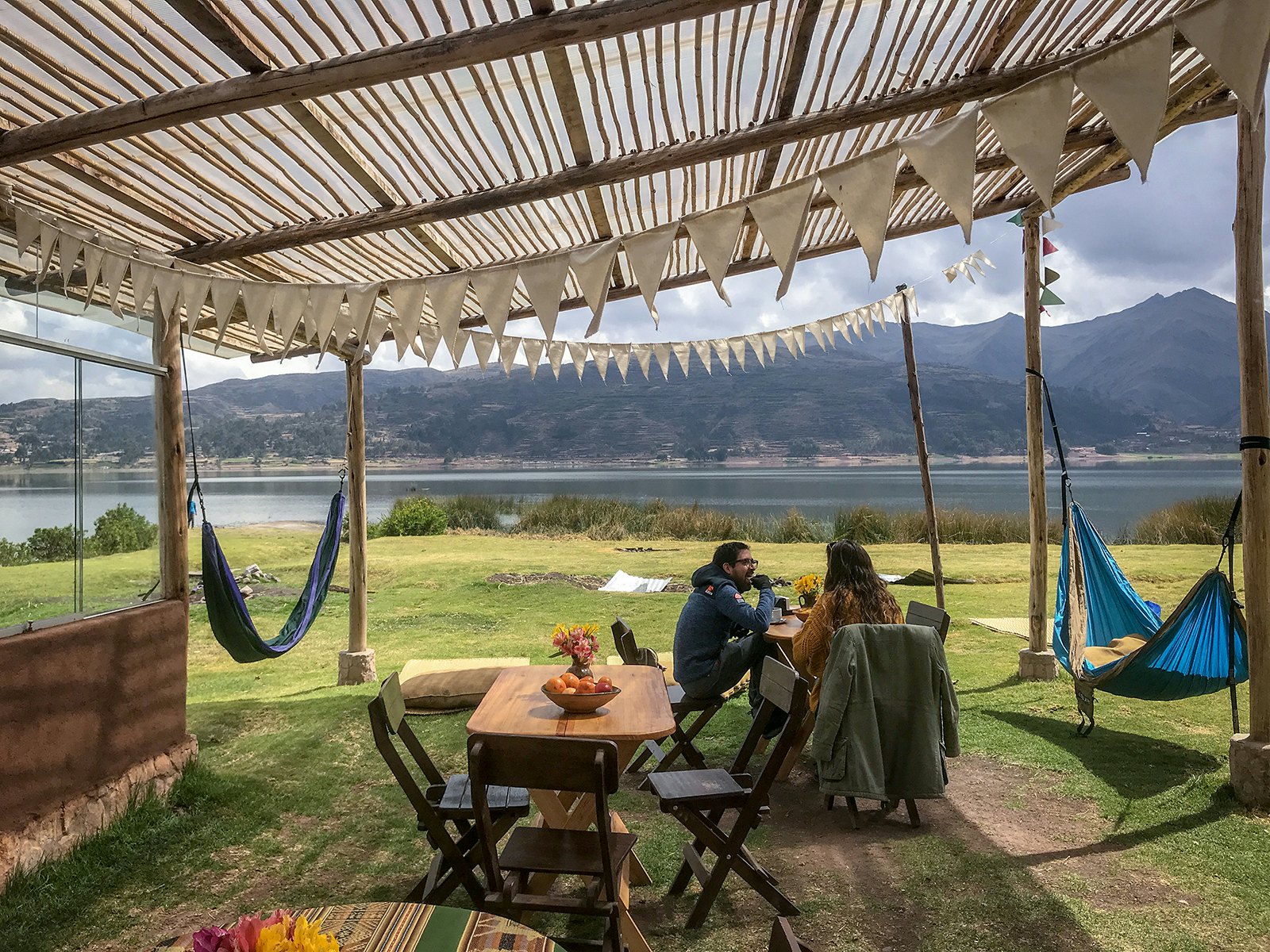 A man and a woman sit at a table in an open-air pavilion, with hammocks strung up on either side and a lagoon in the background