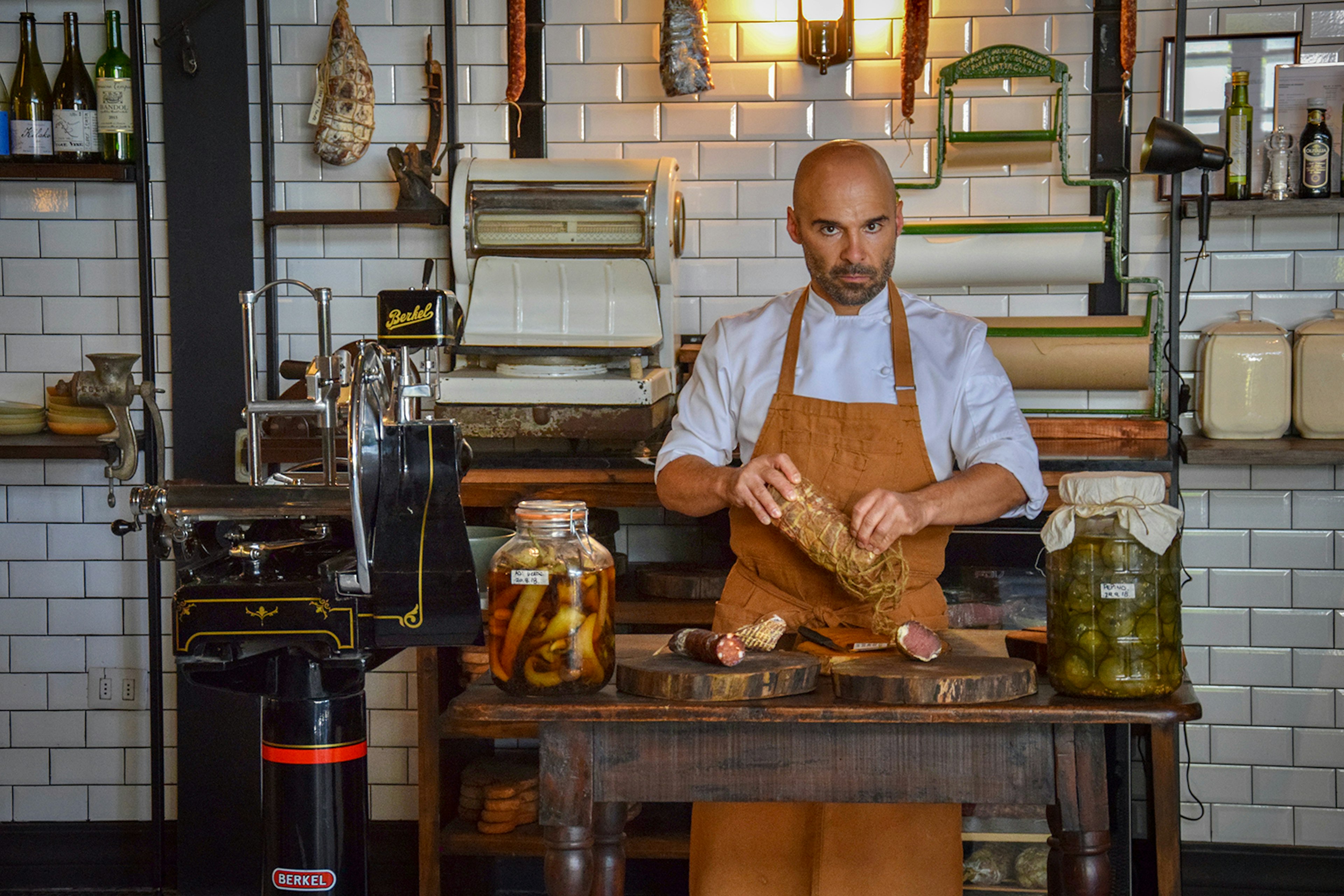 A man wearing a tan apron looks into the camera while holding dried meat at a charcuterie station in Santiago, Chile