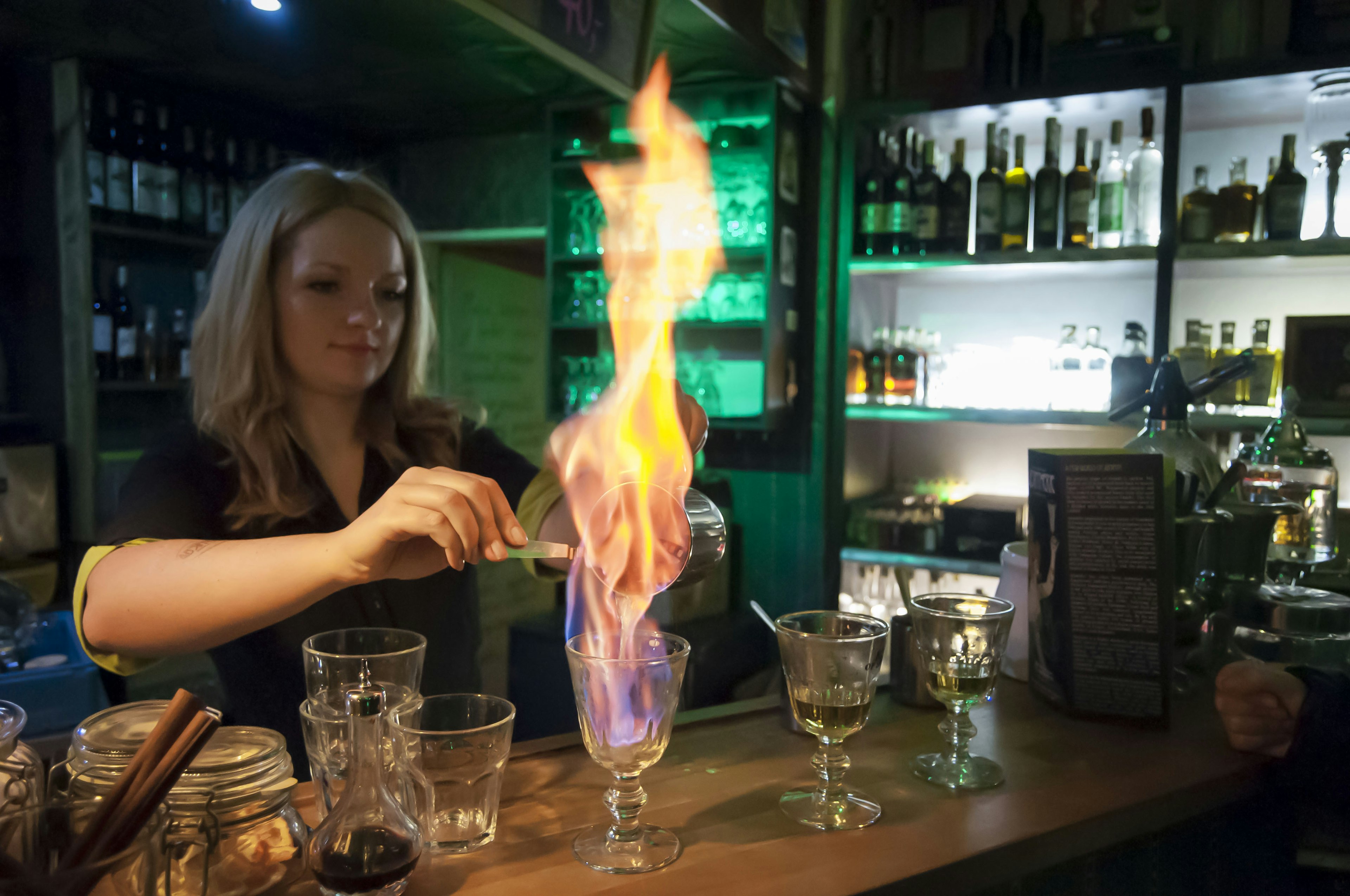 A fairly large flame shoots from a glass as a bartender pours absinthe into it.