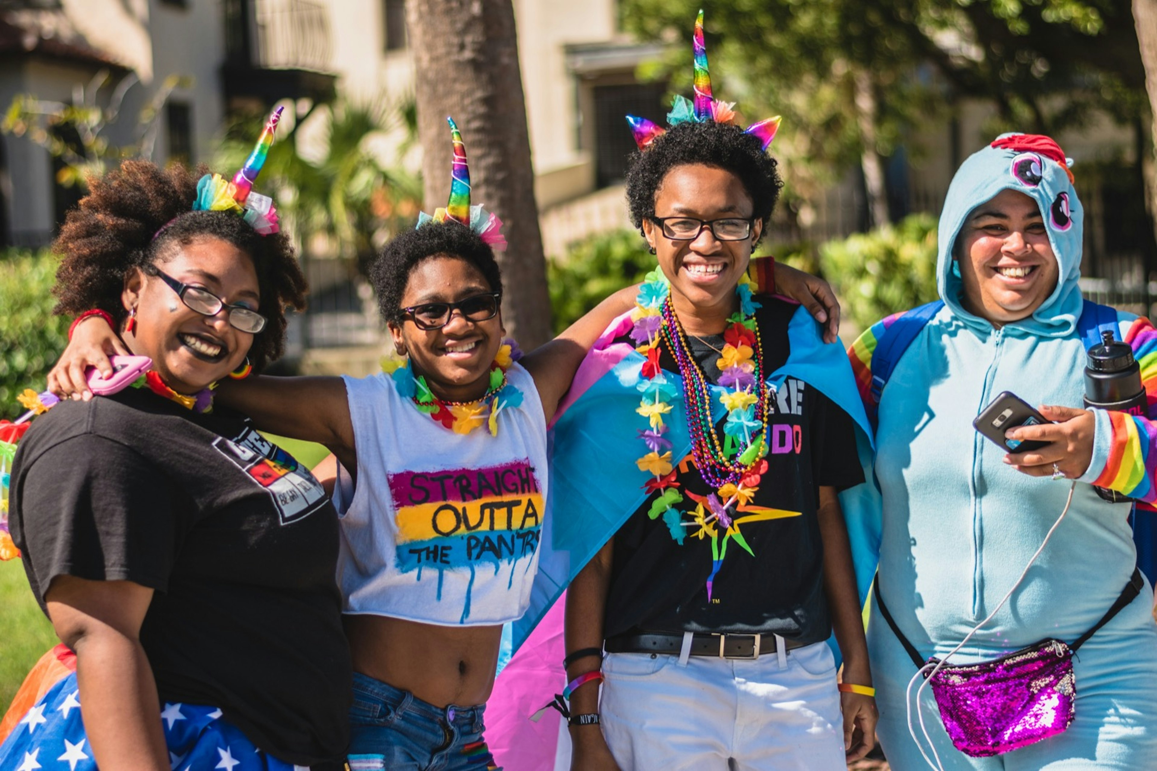 A group of four people decked out in unicorn horns, beads and plastic flowers. One person is wearing a unicorn onesie and another is wearing a Wonder Woman skirt. NYC WorldPride is hosting CosPlay and Pride in June.