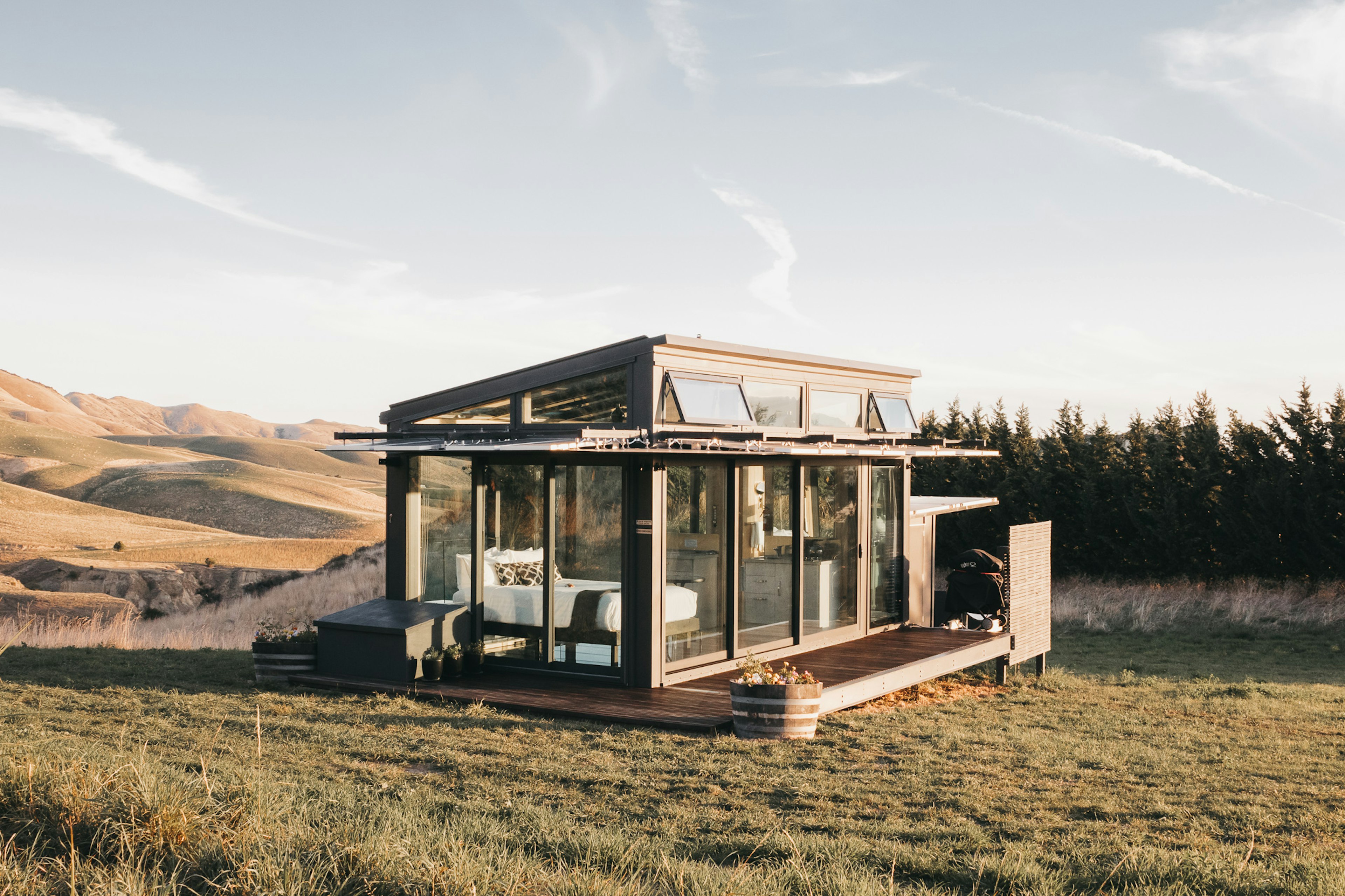 A simple wood-and-glass chalet stands on grass with rolling hills beyond.