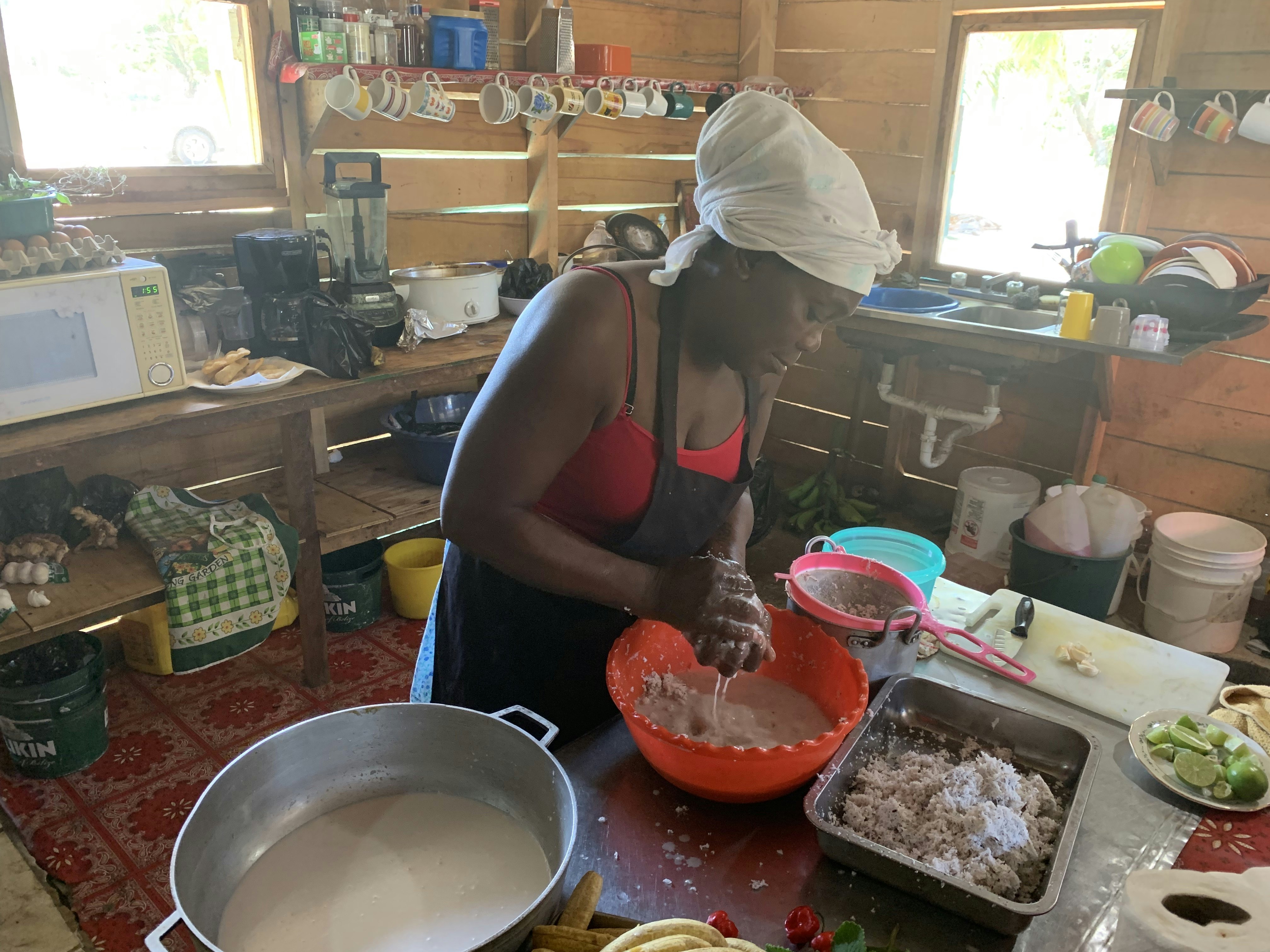 A woman wearing a red top, black apron and white headscarf squeezes milk into a plastic bowl from coconut shavings