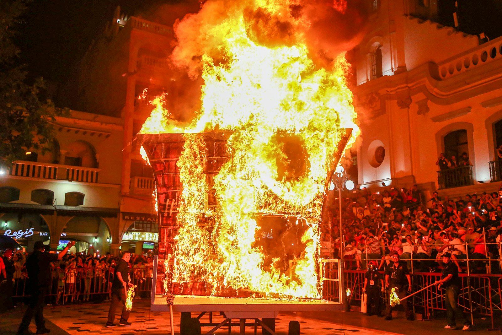 A large effigy is completely engulfed in flames as a large group of people look on