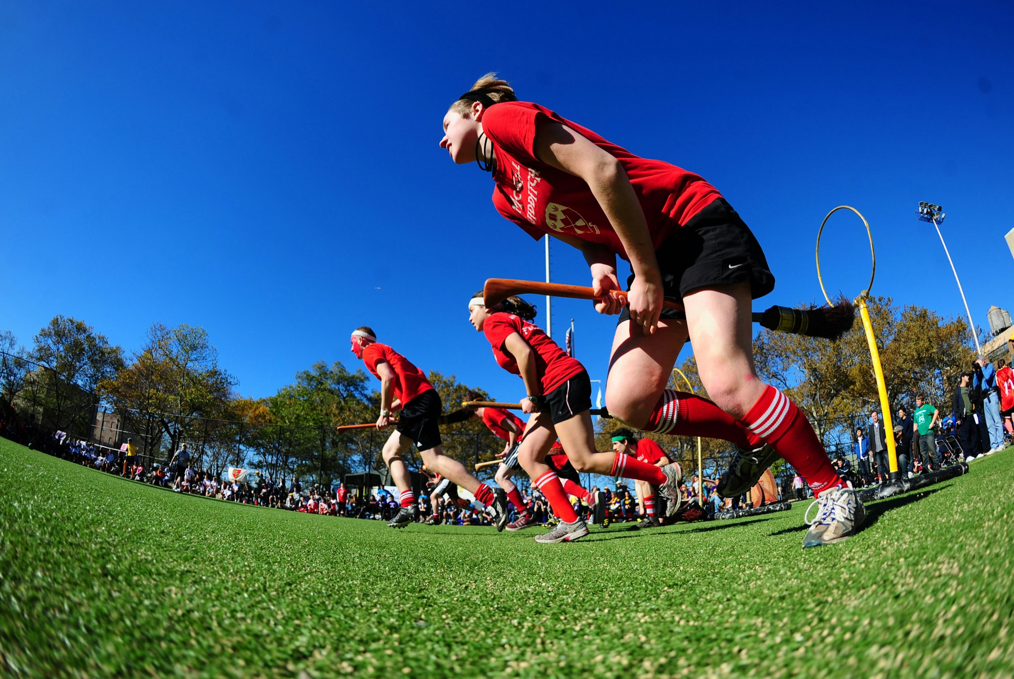 Players in red jerseys run across a field with broomsticks between their legs; Unique sporting events