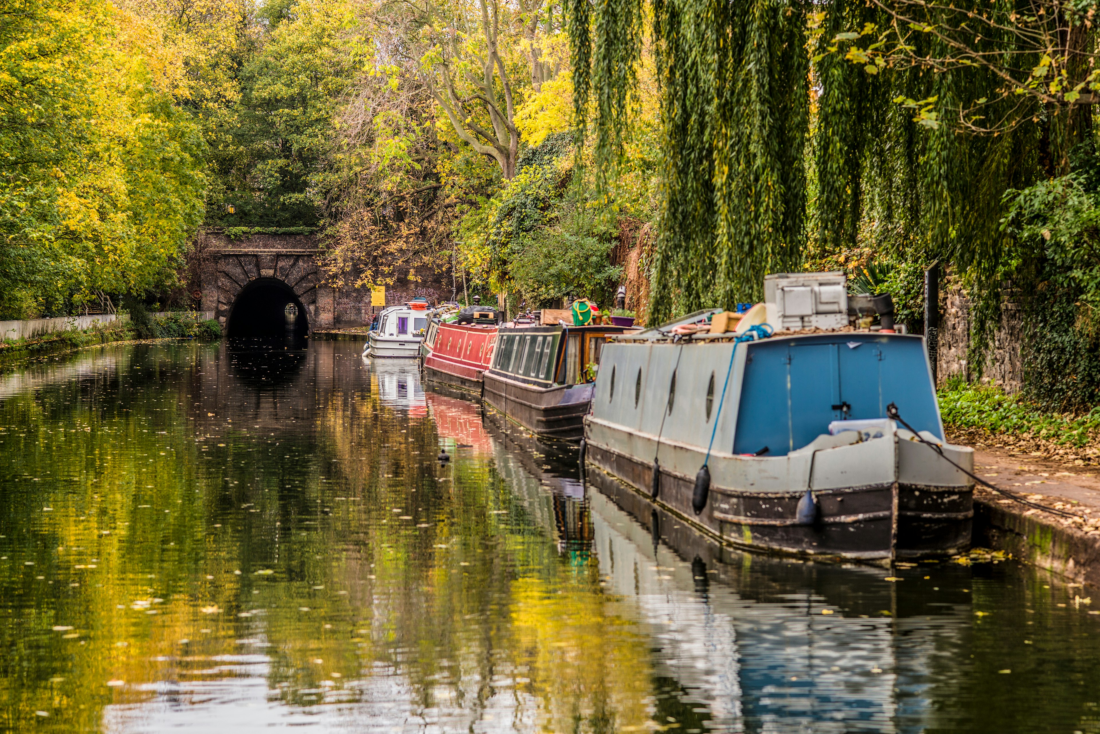 Canal boats docked alongside a narrow canal, with fall colors on the banks and a tunnel in the background