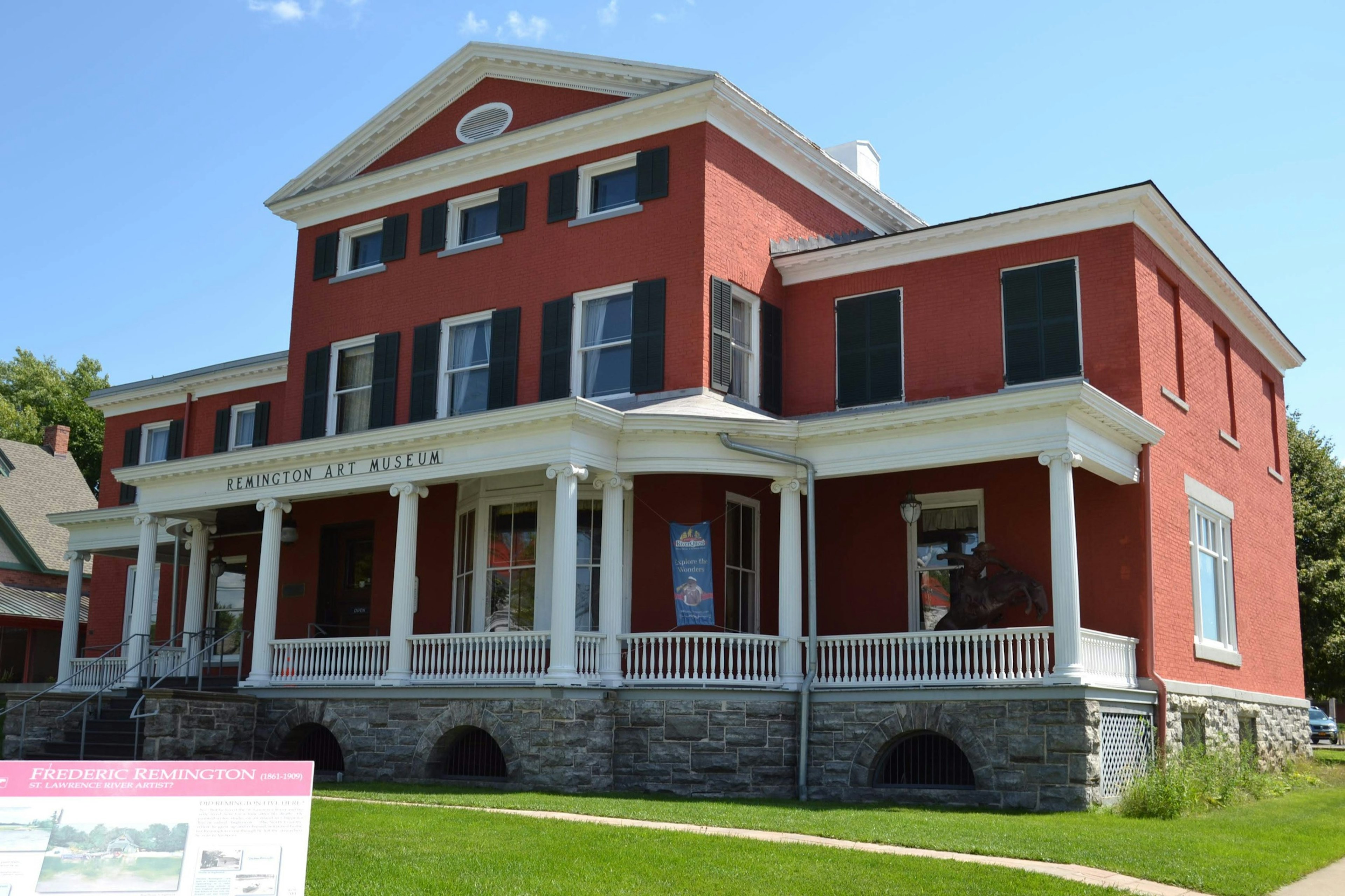 A red house with a large white-pillared front porch is seen on a sunny day with a sign out front proclaiming it the Frederic Remington museum; Where to see Remington in America