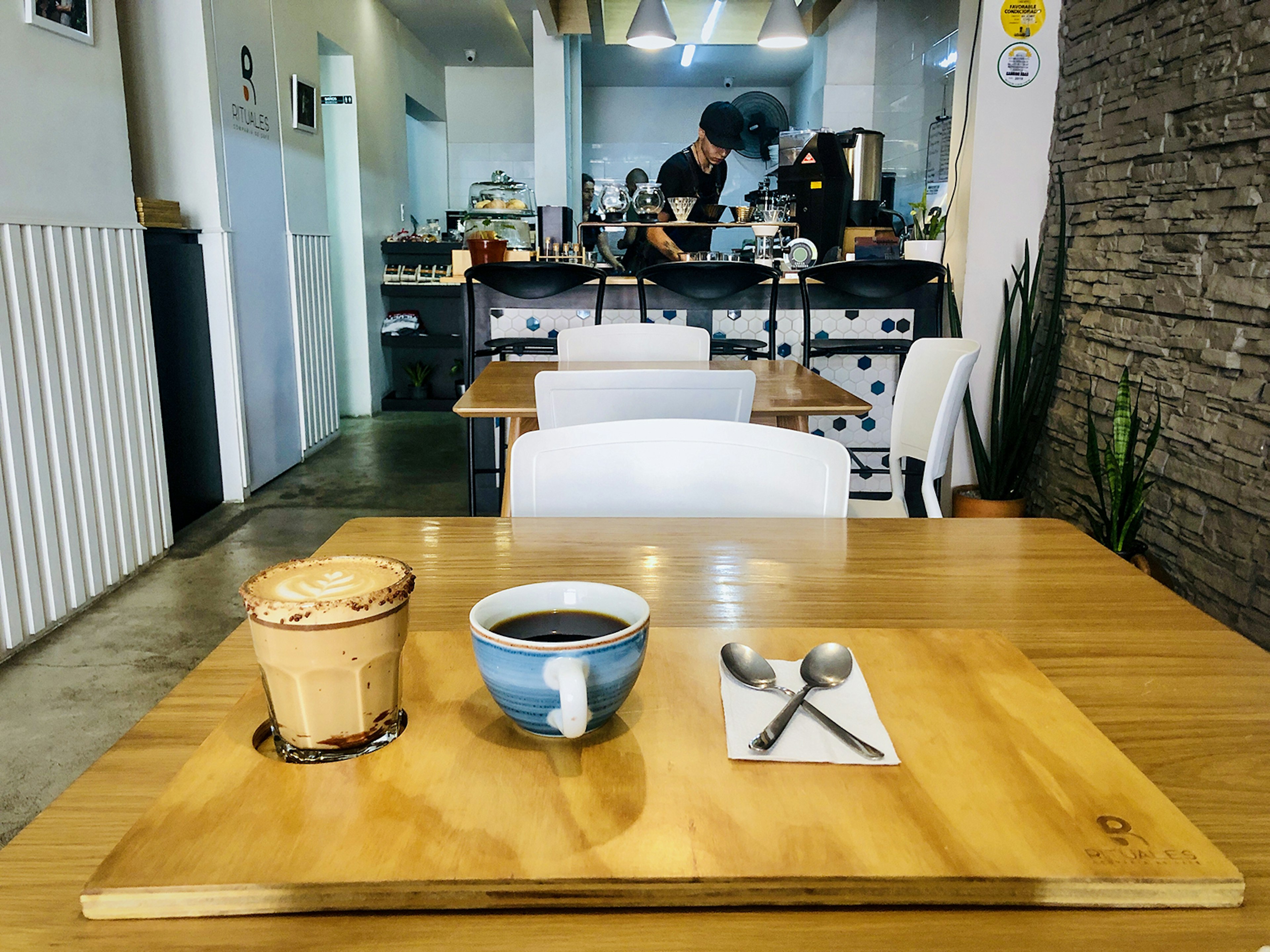 View from a wooden table set with a coffee presentation that includes a latte, a cup of black coffee and spoons crossed on a napkin. A person prepares coffee at a bar in the background. Medellín, Colombia.