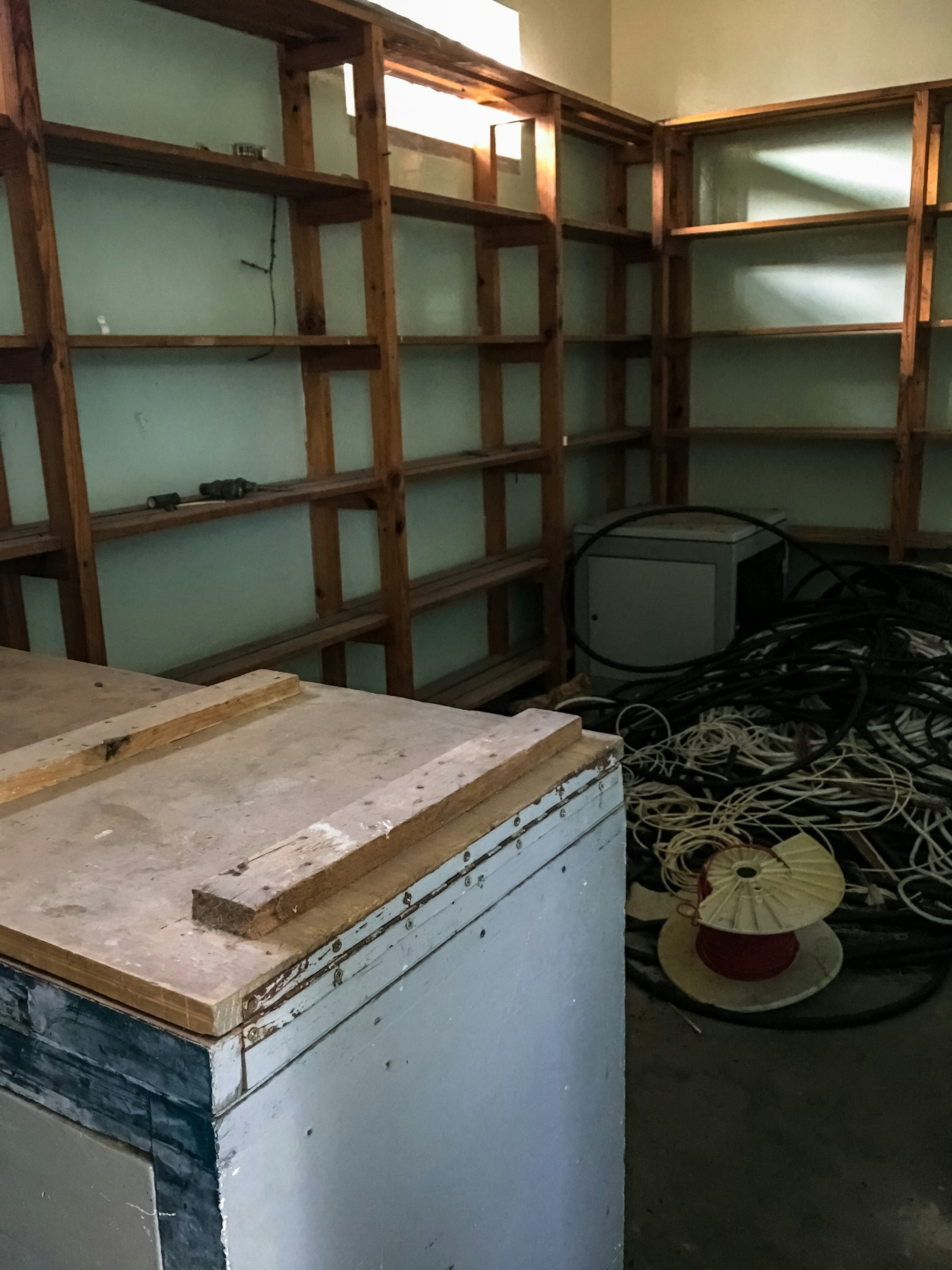 A dishevelled room with the floor covered in old electrical cables; the pale green walls have empty wooden bookshelves on them.