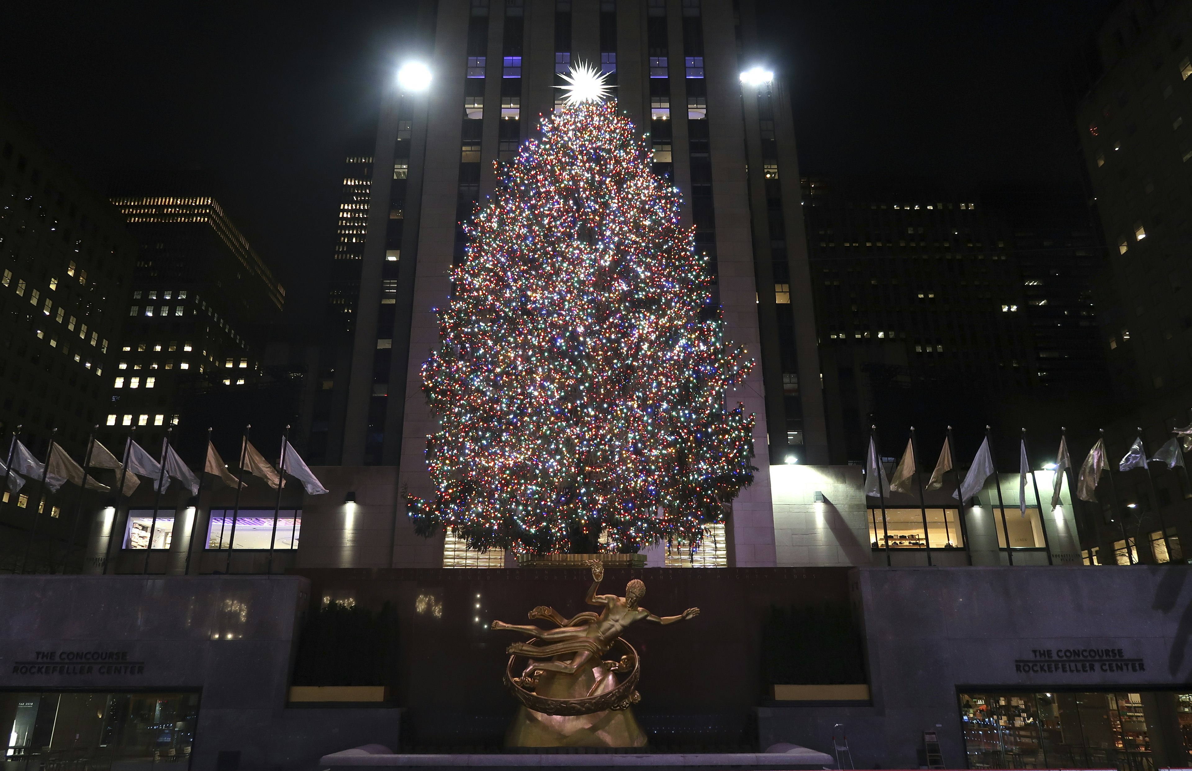 The Rockefeller Center Christmas tree shines brightly in front of the golden Prometheus statue at Rockefeller Center. There's a large bright start at the top of the tree.