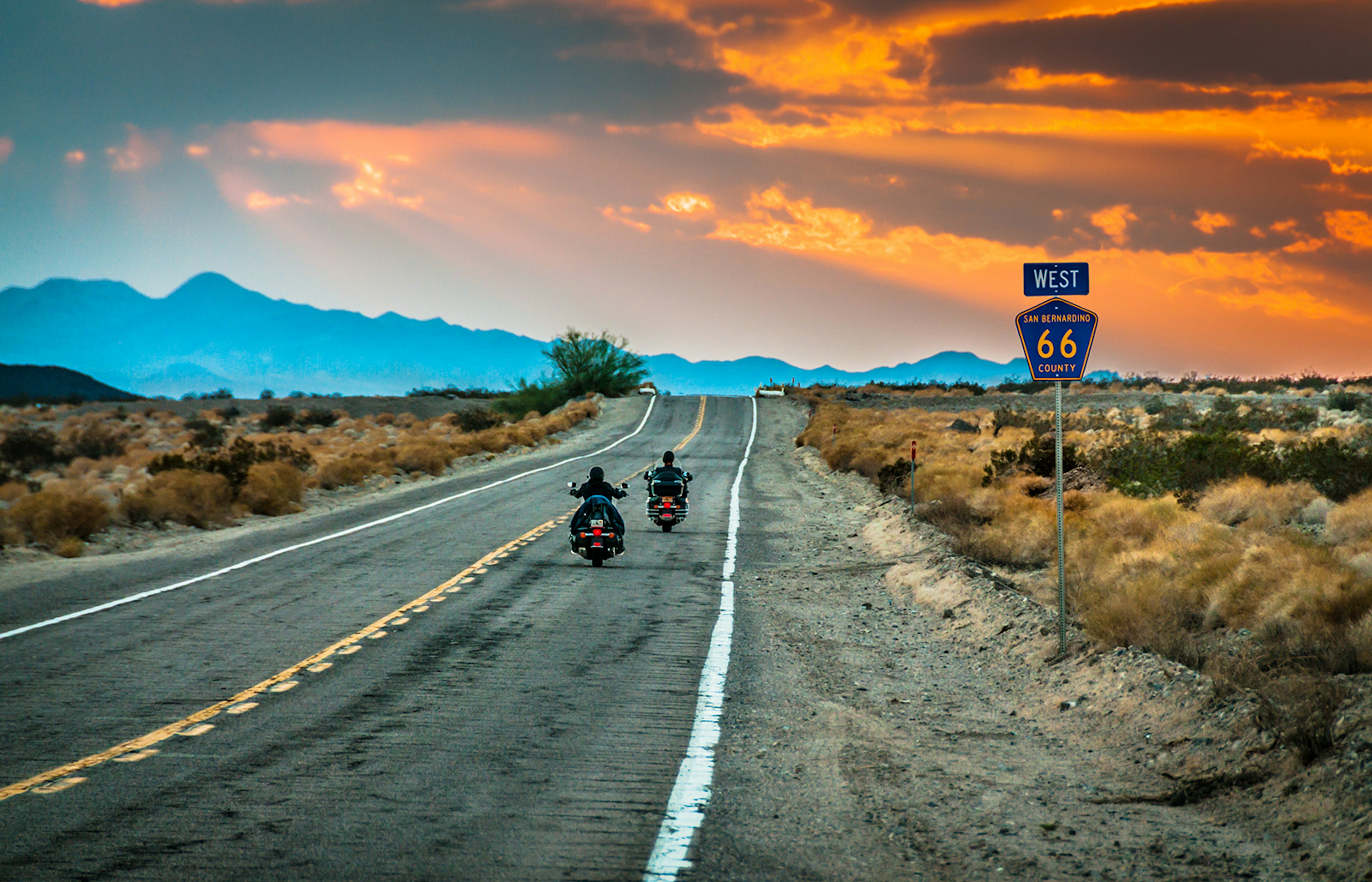 Western US road trip - Two people ride down an empty road on motorcycles at sunset. Mountains are visible in the background, and the sky is orange in hue.