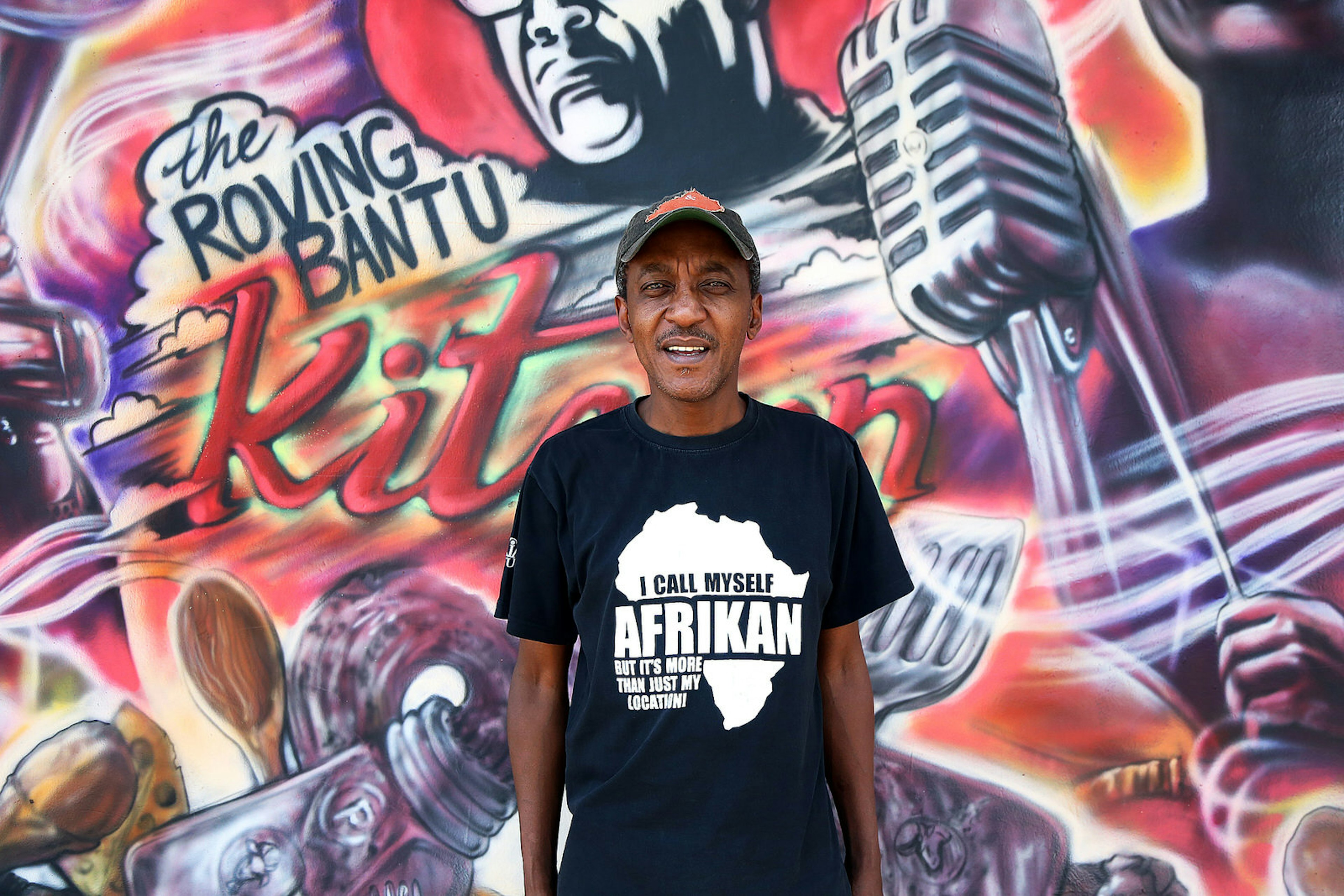 Sifiso Ntuli, stands in front of his restaurant which is covered in a street art mural. He wears a T-shirt that says, 'I all myself Afrikan, but it's more than just my location!' © Heather Mason / ϰϲʿ¼