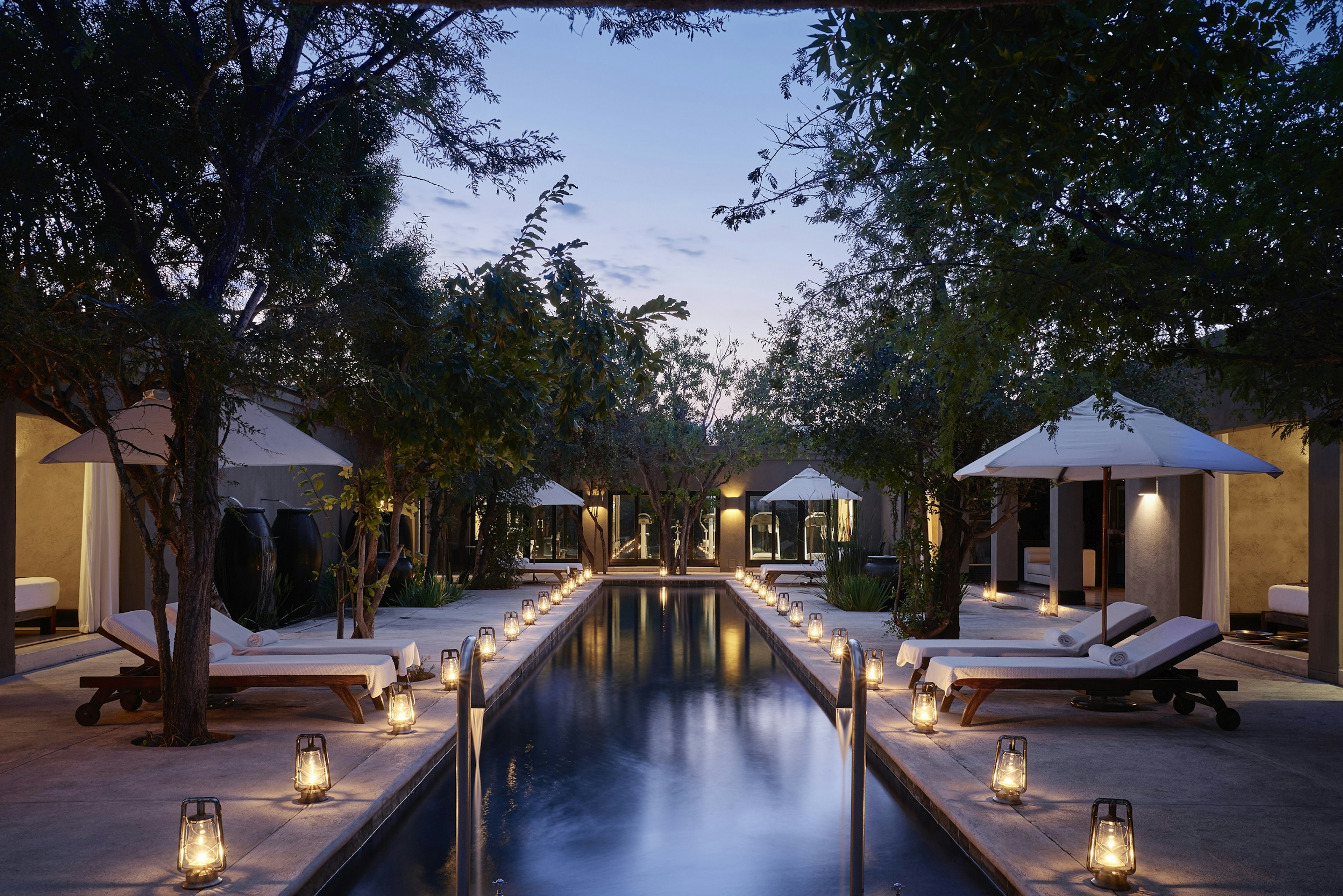 A swimming pool is surrounded by deck chairs and umbrellas at dusk. Lamps line the side of the pool.