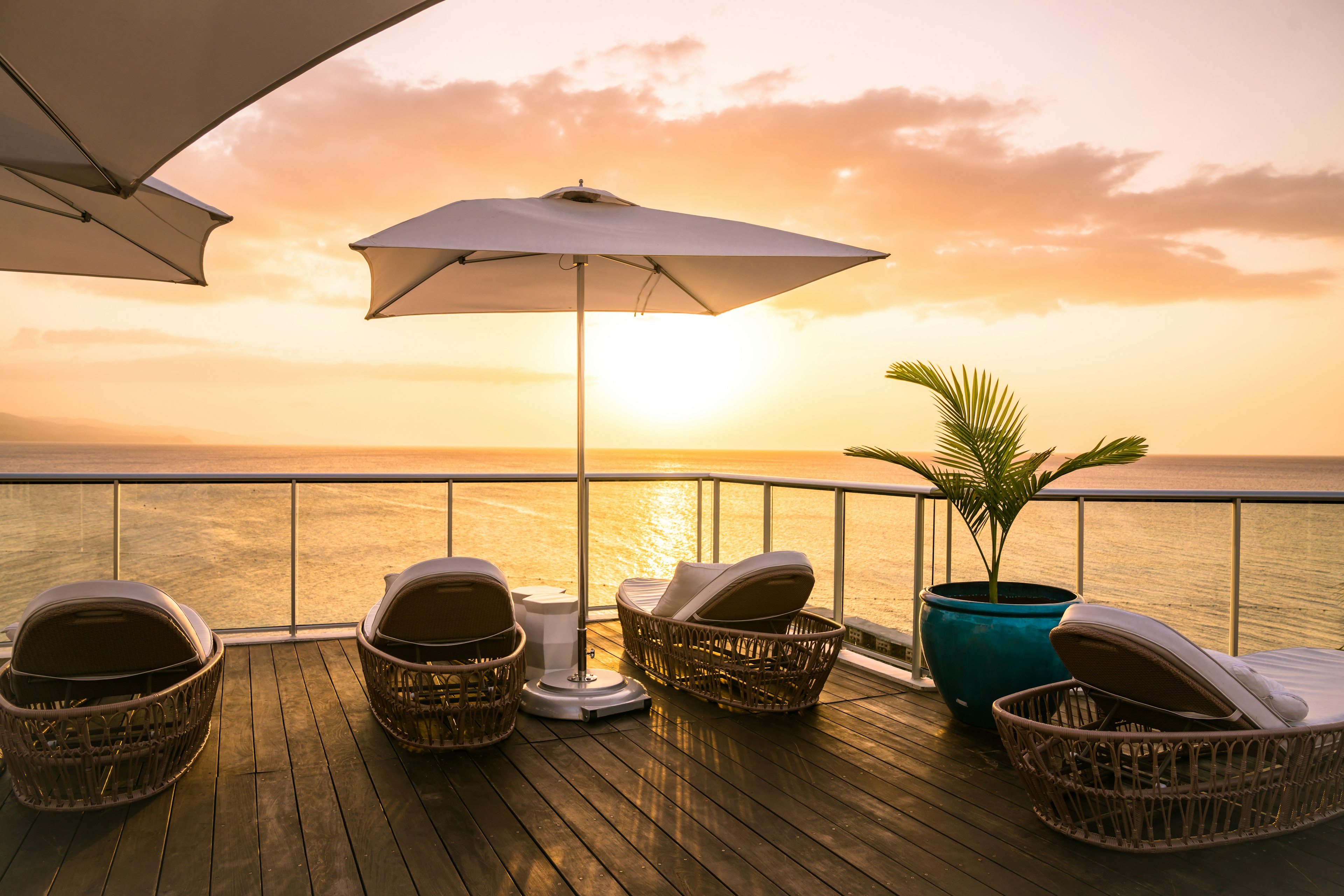 A collection of rattan lounge chairs, a large umbrella and a large potted plant face the ocean as the sun sets on the sky deck area at the S Hotel in Jamaica.