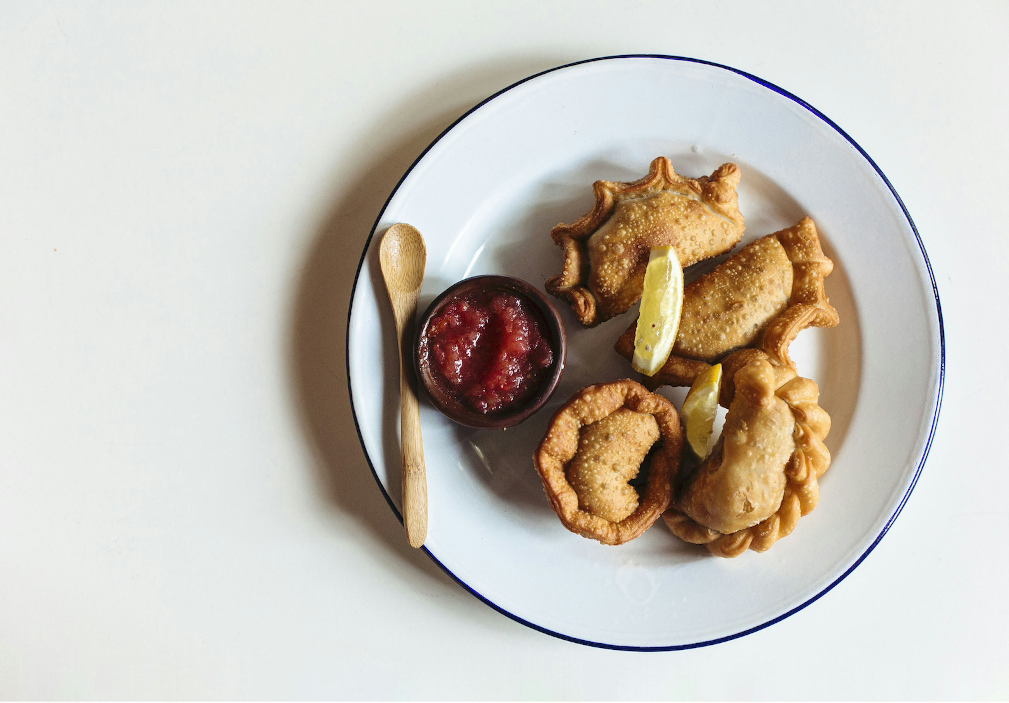 Four perfectly folded empanadas served on a white plate with a ramekin of sauce and a wooden spoon on the left side of the plate