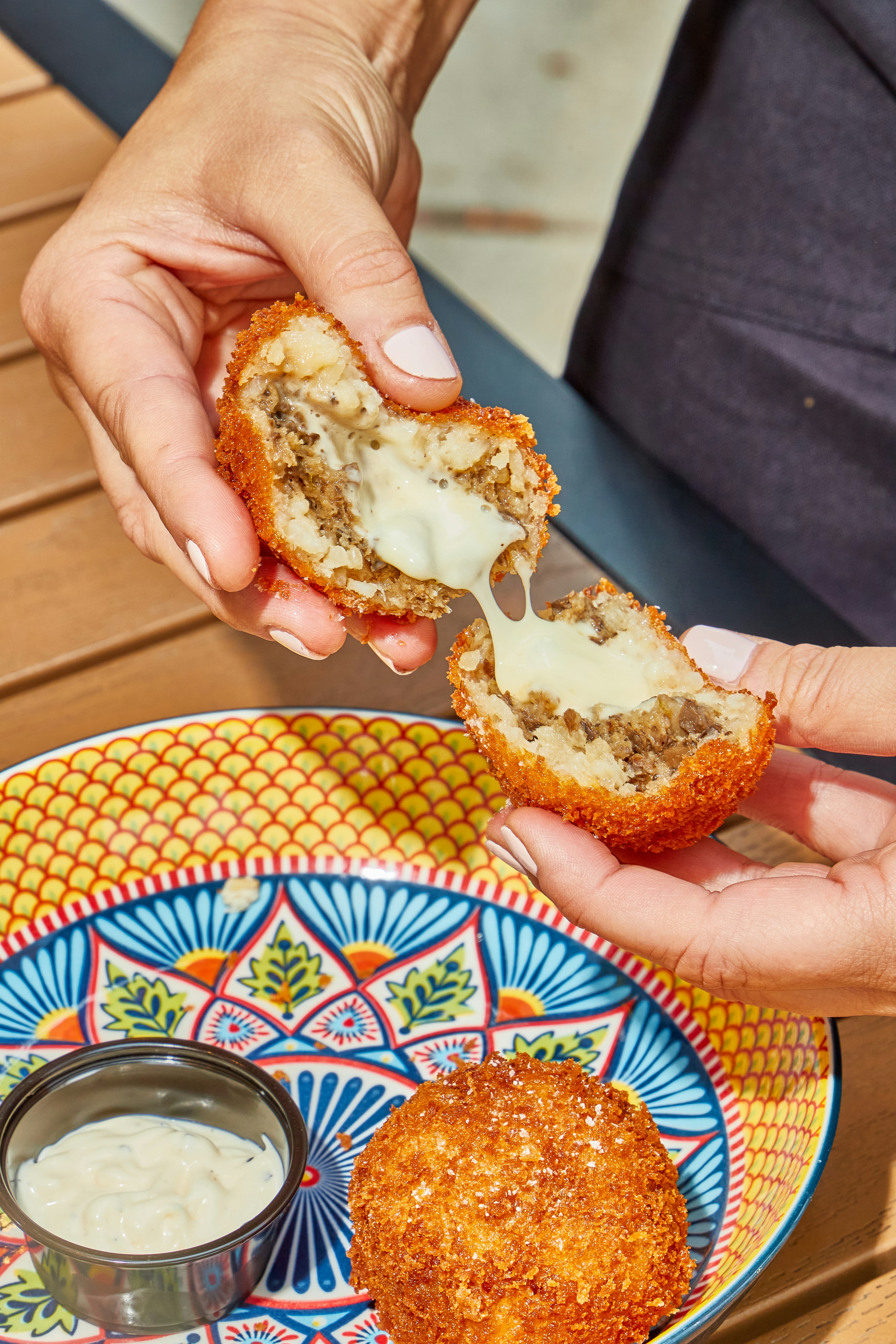 A person pulls apart a crispy arancini ball over a colorfully pattered bowl containing a dip and another arancini