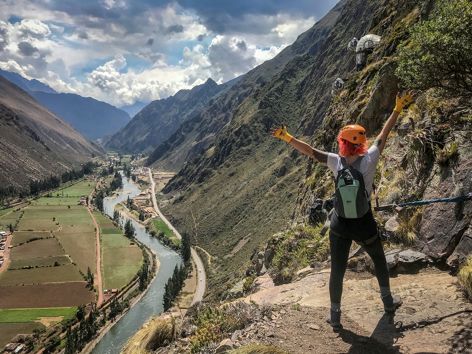 A girl with short orange hair stands on a high rock ledge with her back to camera, gazing out over a river in the Sacred Valley in Peru