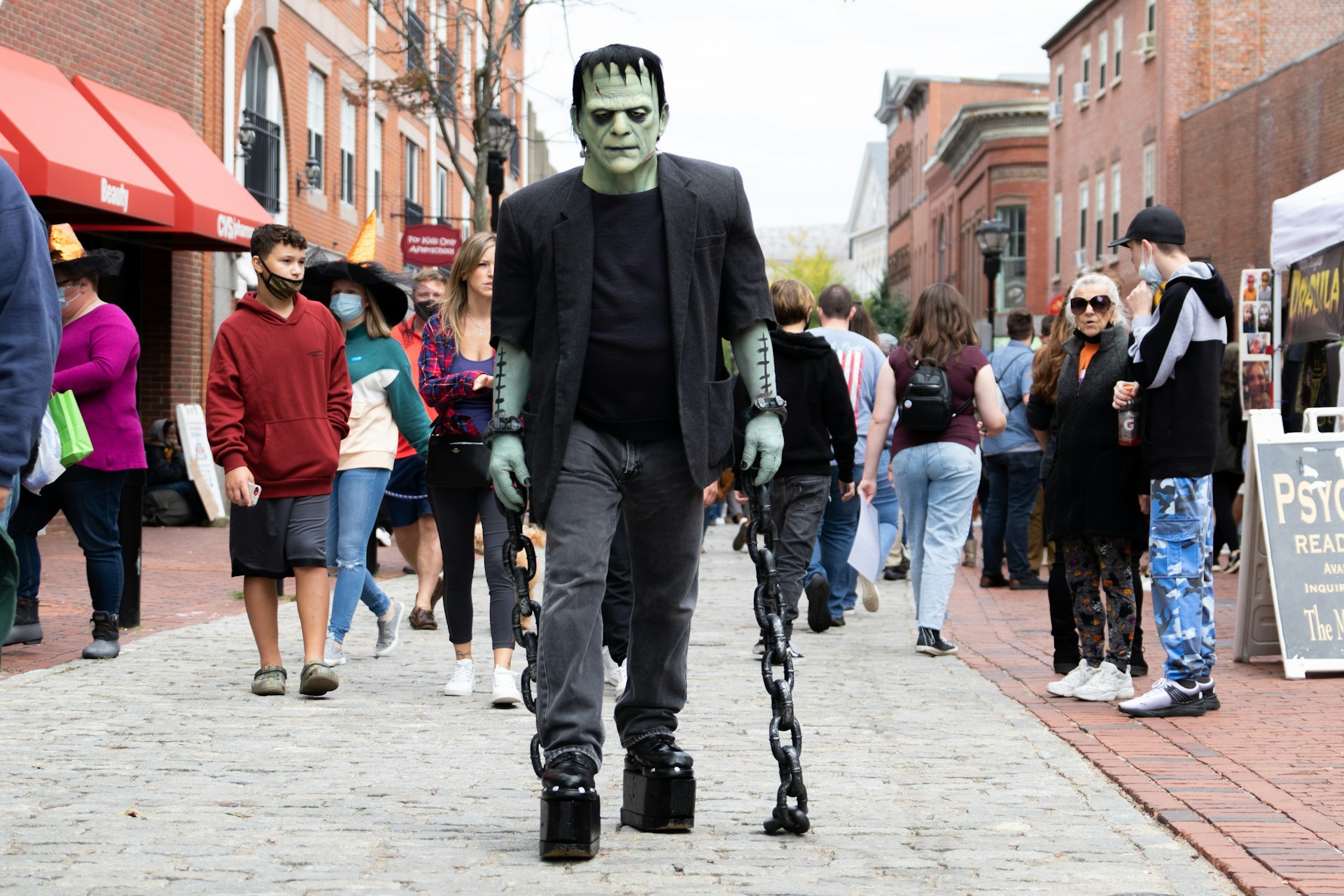 A large monstrous-like figure with white face paint and wearing all black, replicating Frankenstein's monster, walks down a street