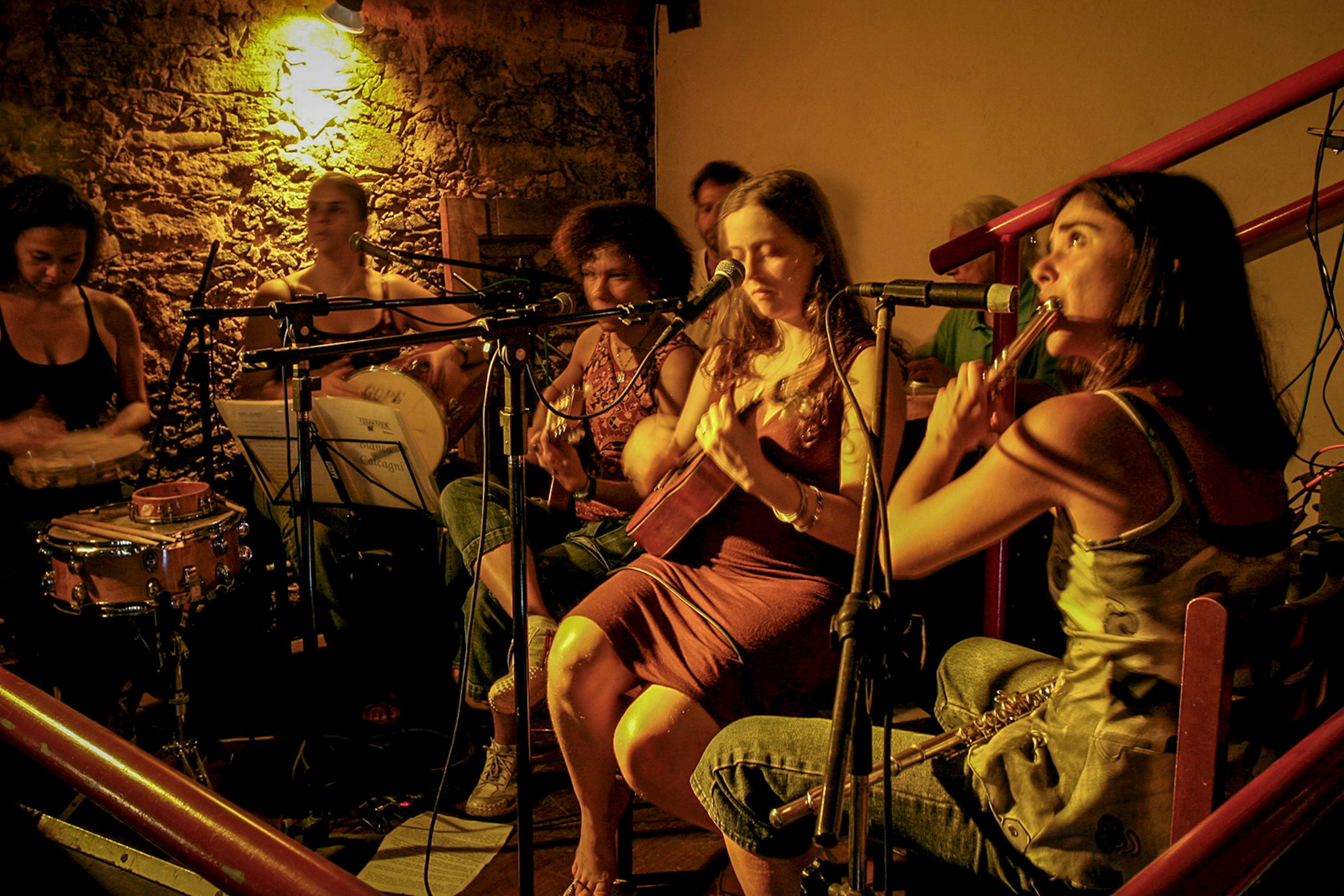 Seven musicians play samba sitting in a semi circle in a warmly lit venue