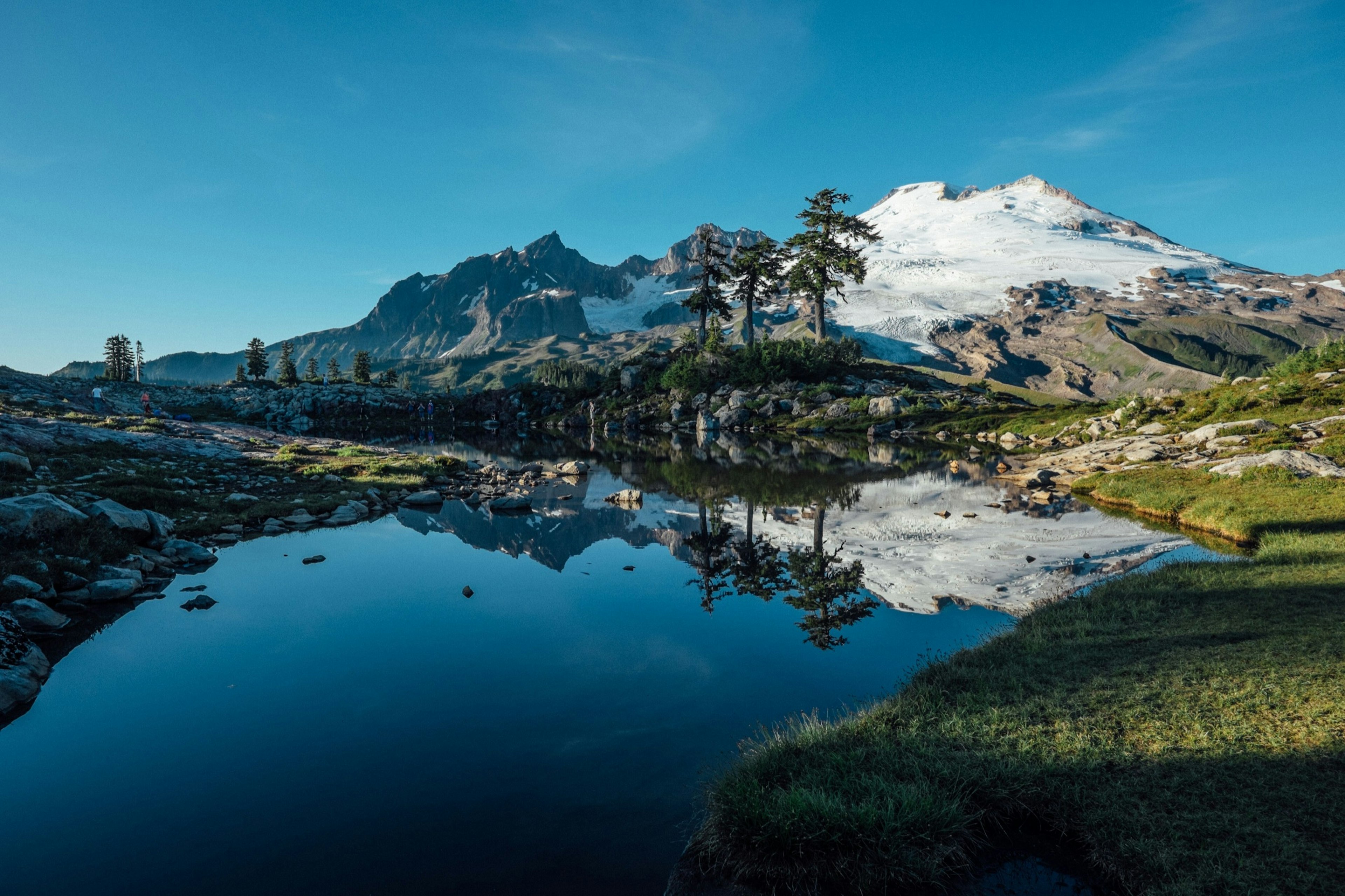 A lovely reflective lake is seen at the foot of a snow capped mountain and a brilliant blue sky among rolling green hills in Washington