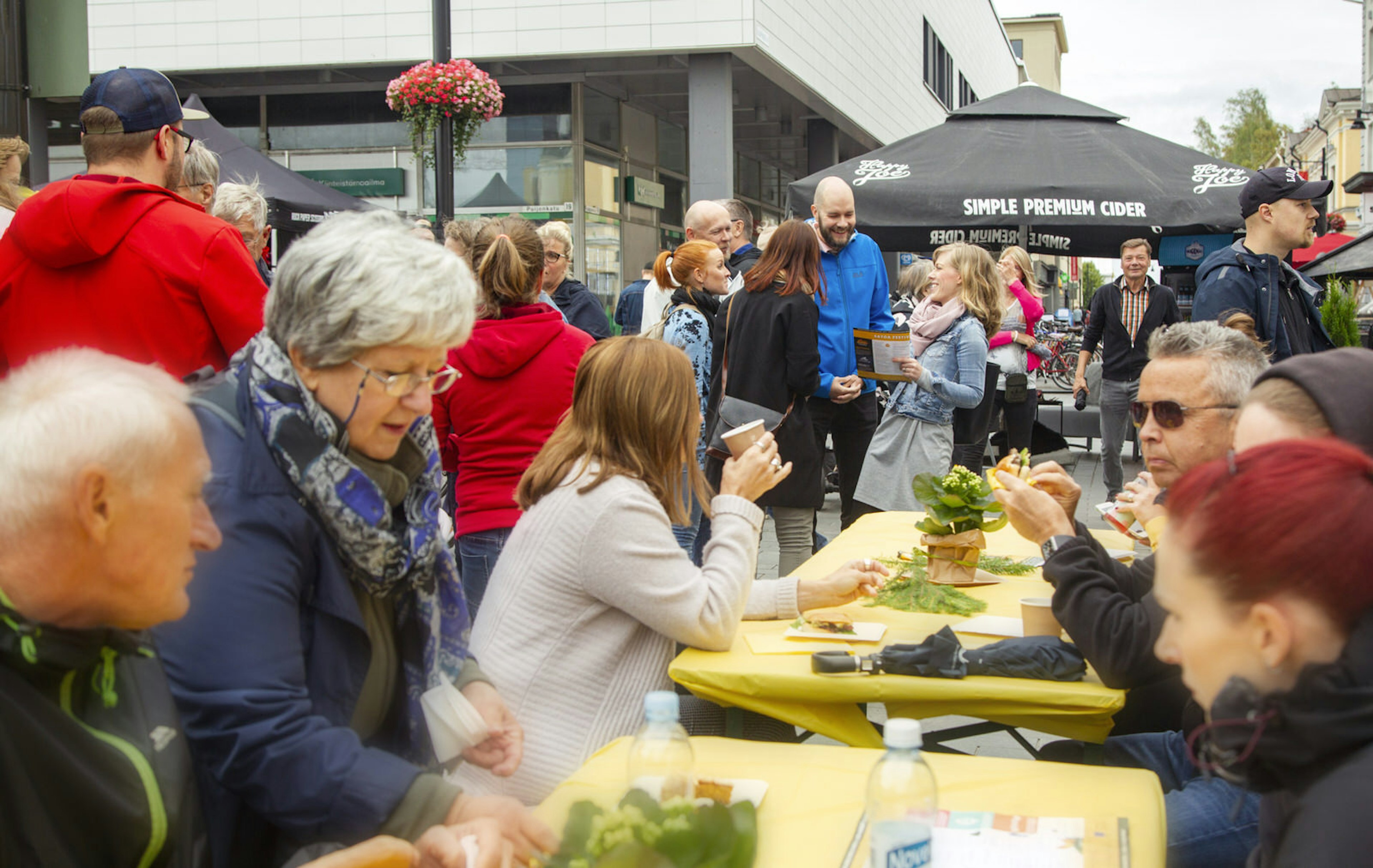 Several people seated at yellow tables, outdoors, eating and chatting.