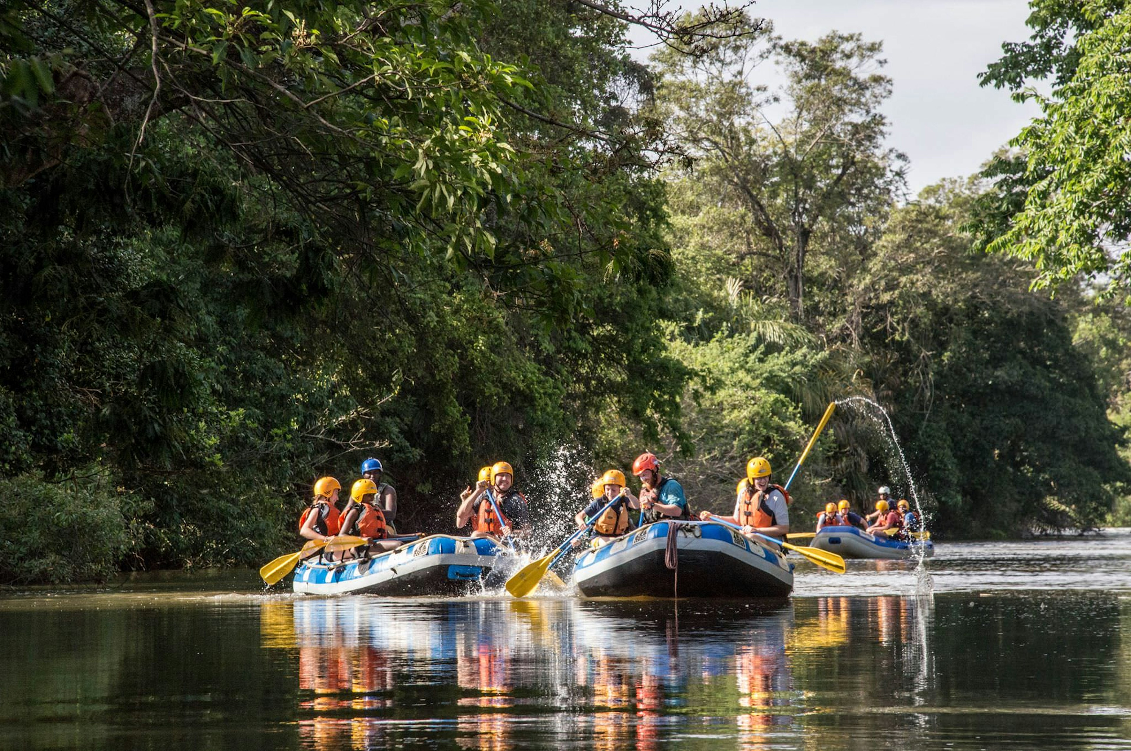 Three rafts, each with five people, floating along a calm river; some of the rafters are splashing water with their paddles.