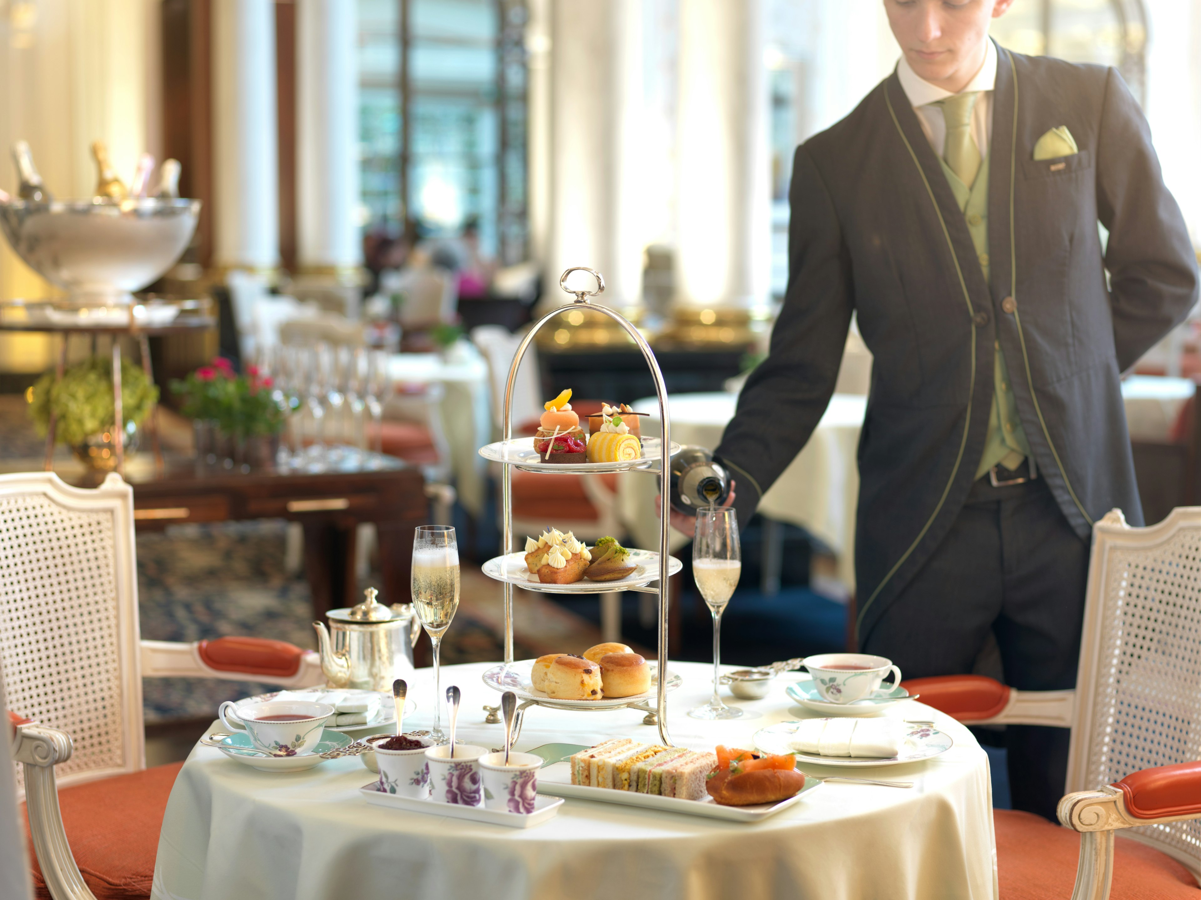 A smartly dressed waiter pours champagne into glasses at a table laden with sandwiches, a cake platter and cups with a floral pattern.