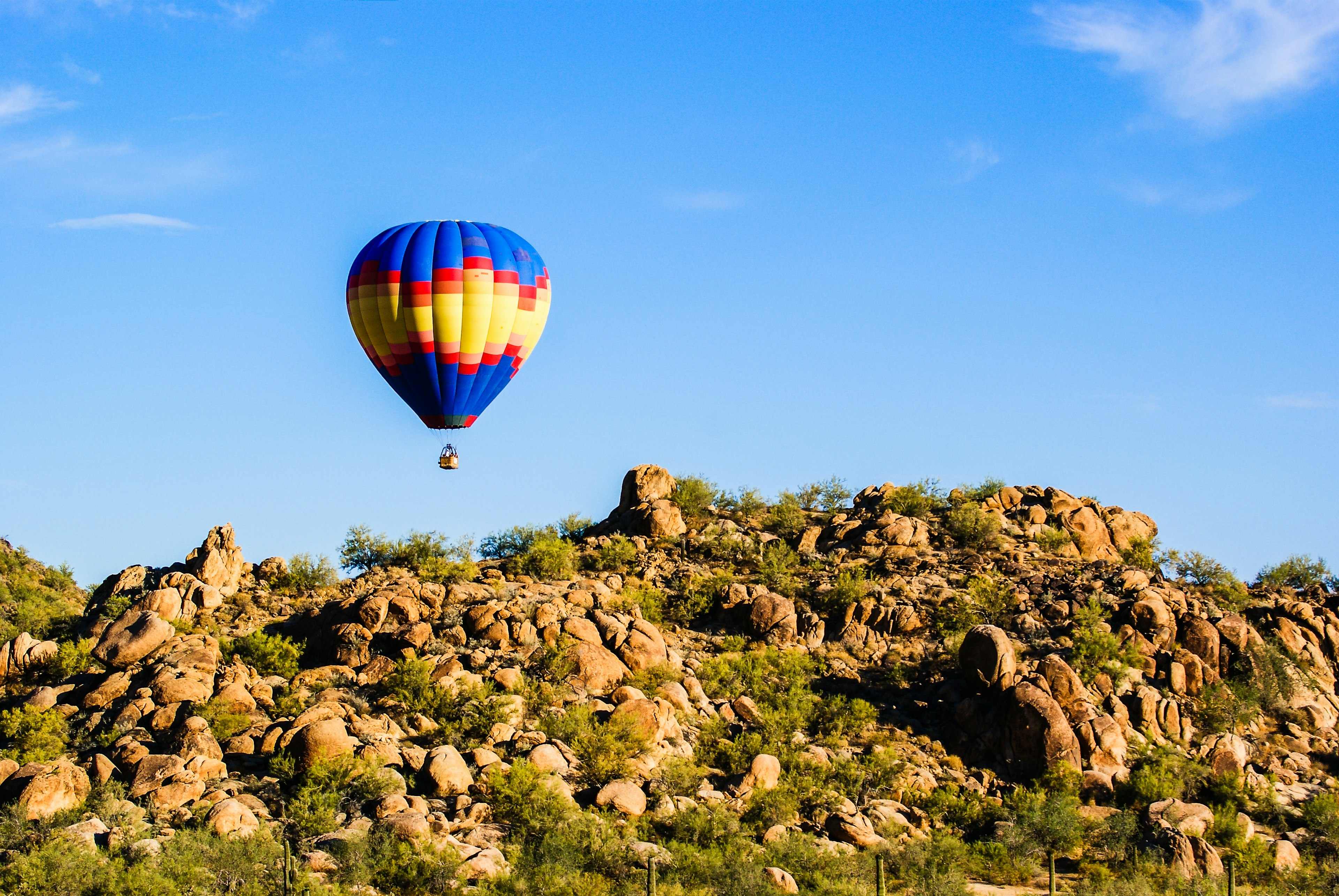 A blue, yellow and red hot-air ballon soars above a rocky hill covered with chunks with greenery; bachelorette party