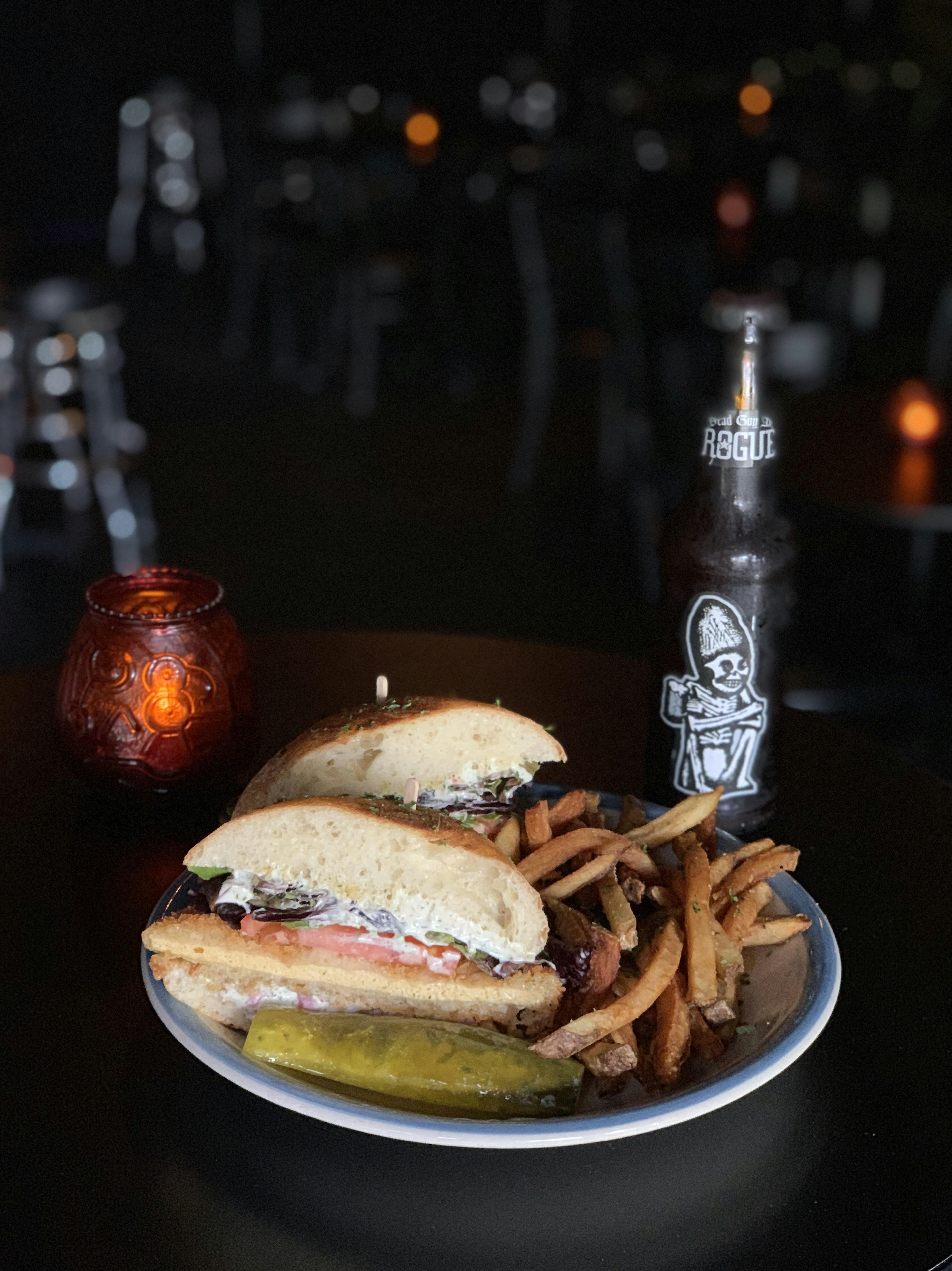 A plate filled with fries, a vegan sandwich and a pickle sits on wooden bar next to a beer bottle and a glass candle holder; Seattle vegan restaurants