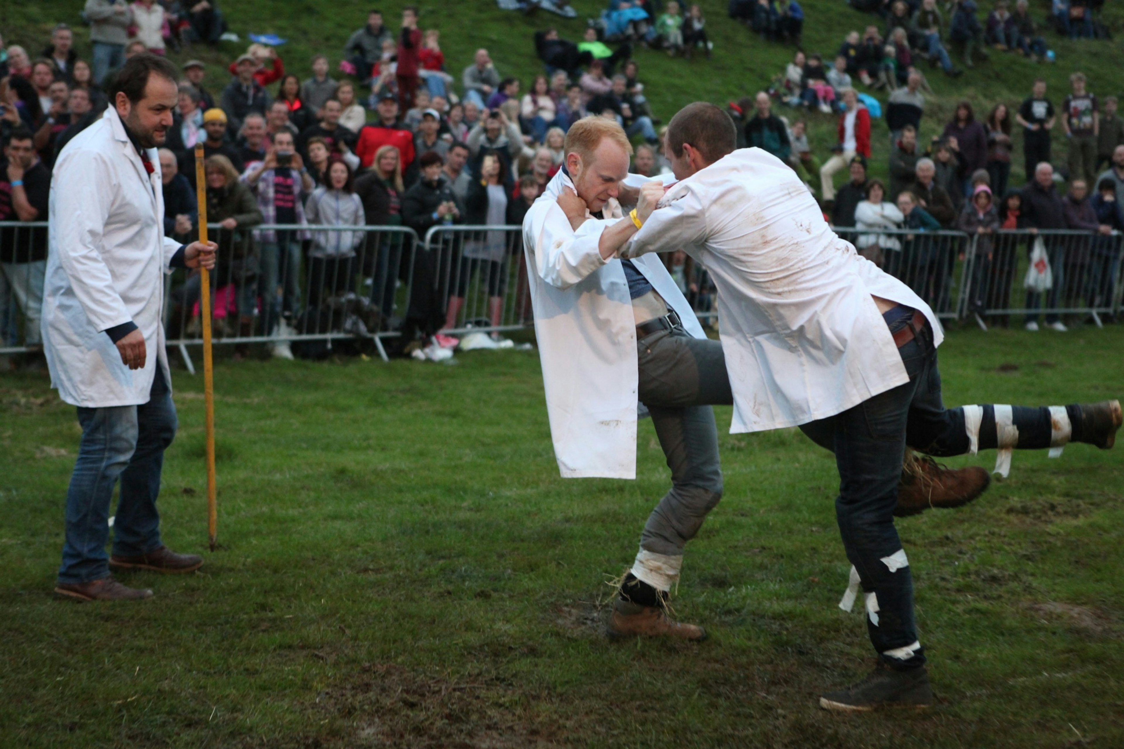 Two men are tangled up while trying to kick each others' shins under the watchful eye of a referee and observers; unique sporting events