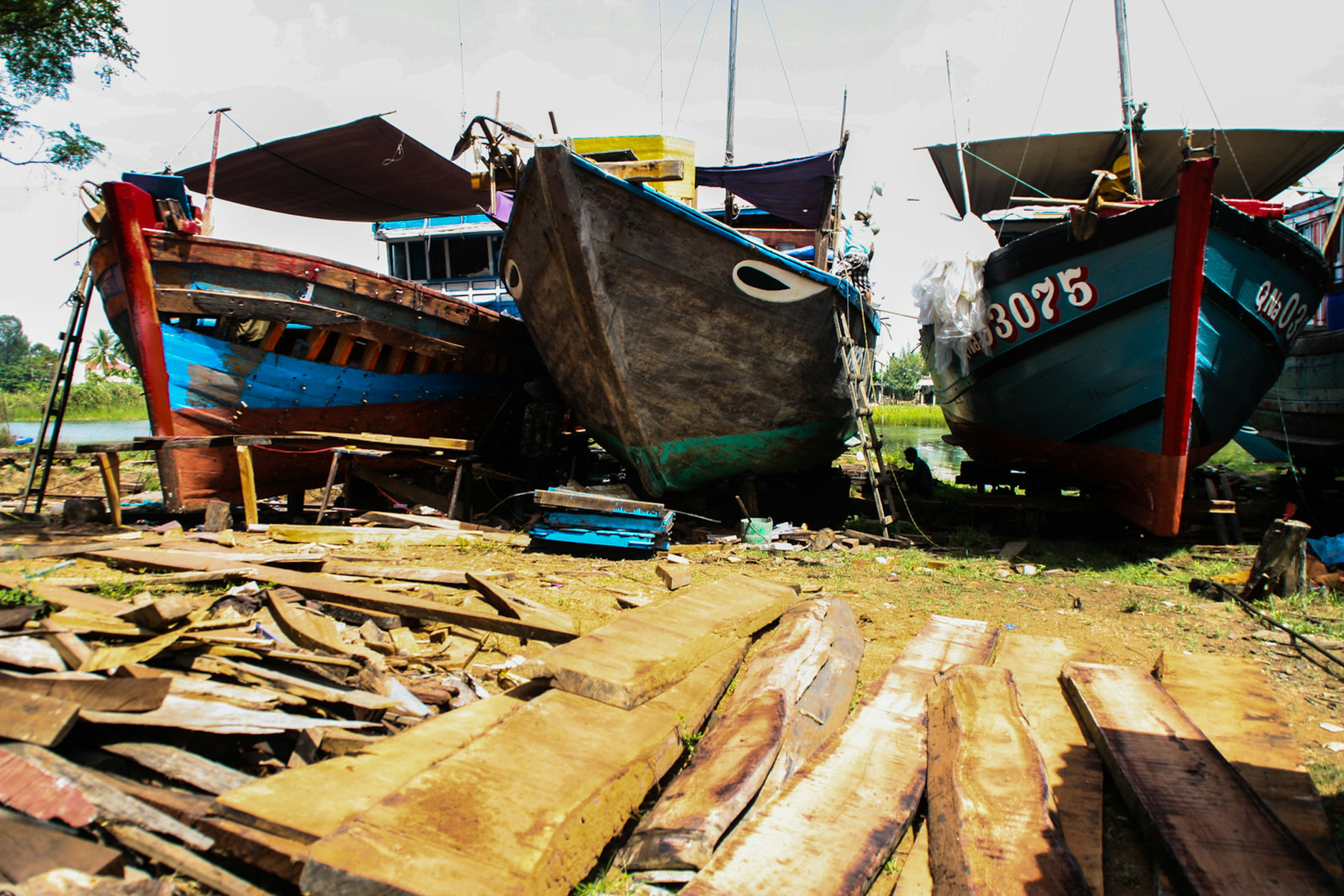 Three wooden ships in various states are lined up on the shoreline, with planks of wood on the ground in front of them