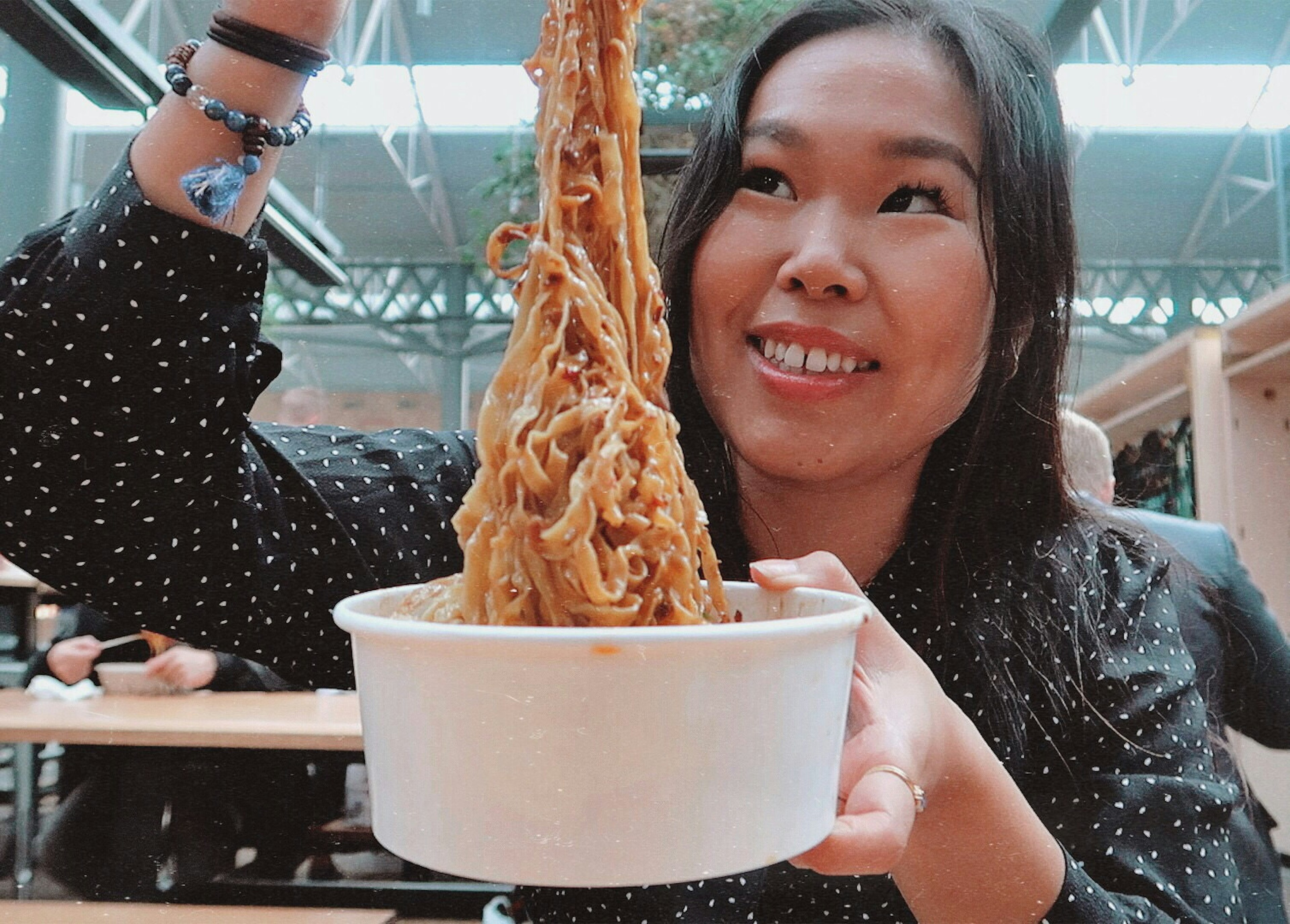 Shu Shi Lin holding up a white paper tub and lifting some very saucy noodles out of it with chopsticks. Shu is looking up at her hand and smiling.