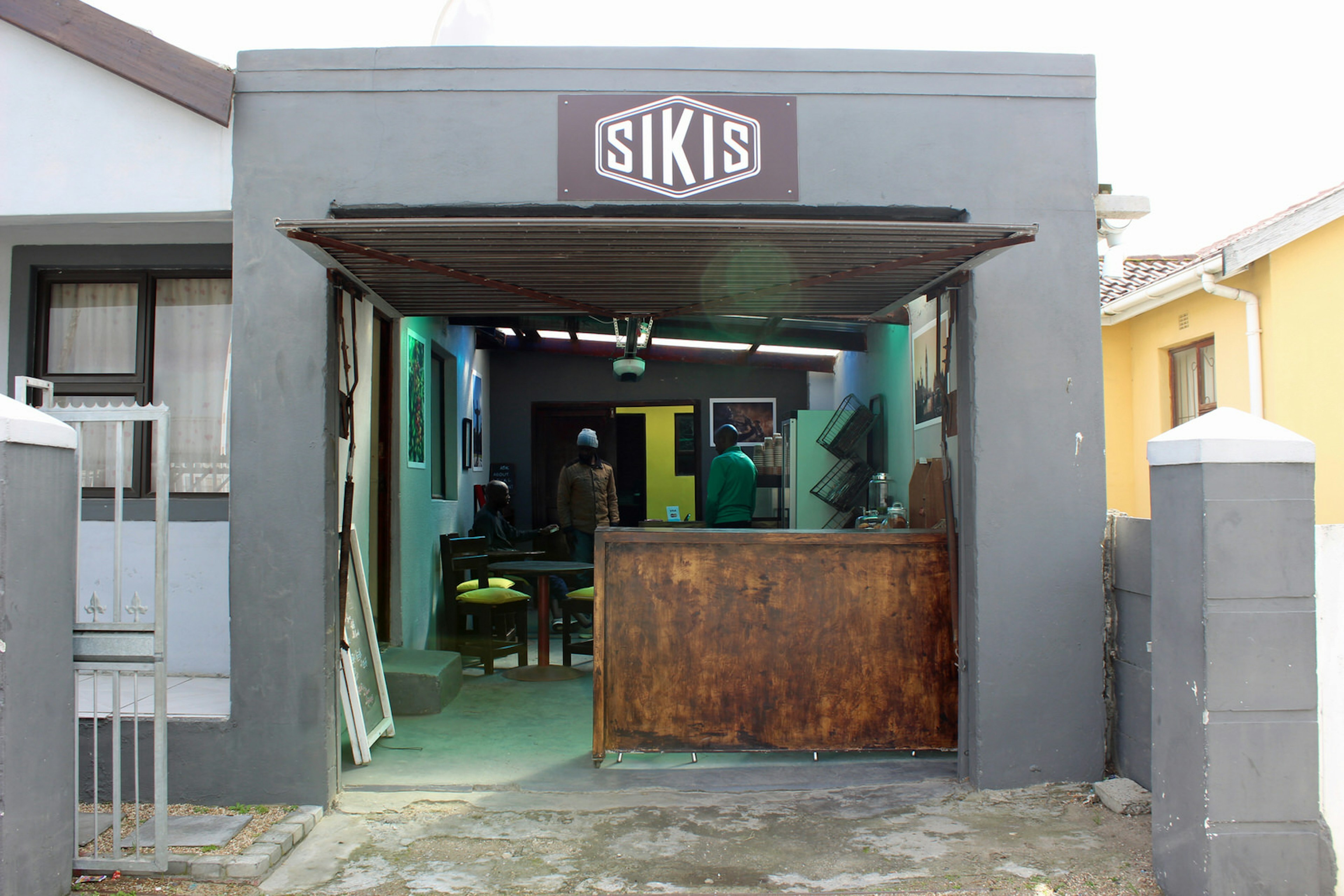 Behind an open garage door in a simple, dark grey box-like exterior is a simple cafe with a wooden bar