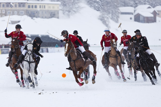 A orange ball hovers above the air as a man holding a wooden mallet prepares to strike it while on horseback. There are a group of other men chasing behind him all on horses during this snow polo match in Lake St. Mortiz