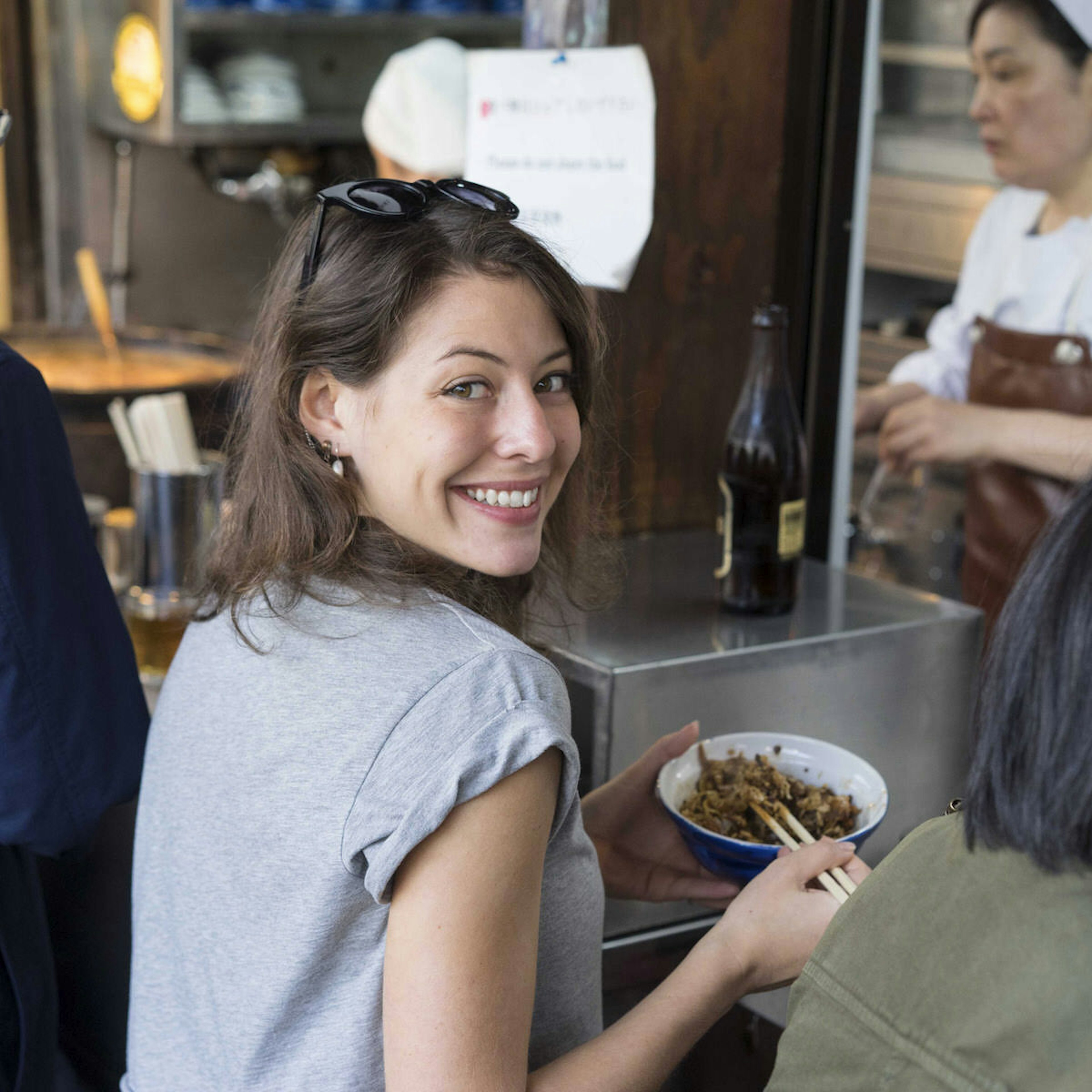 Sofia Levin looks over her shoulder at the camera while eating from a blue bowl with chopsticks. She is wearing a grey t-shirt and has a pair of sunglasses on her head.