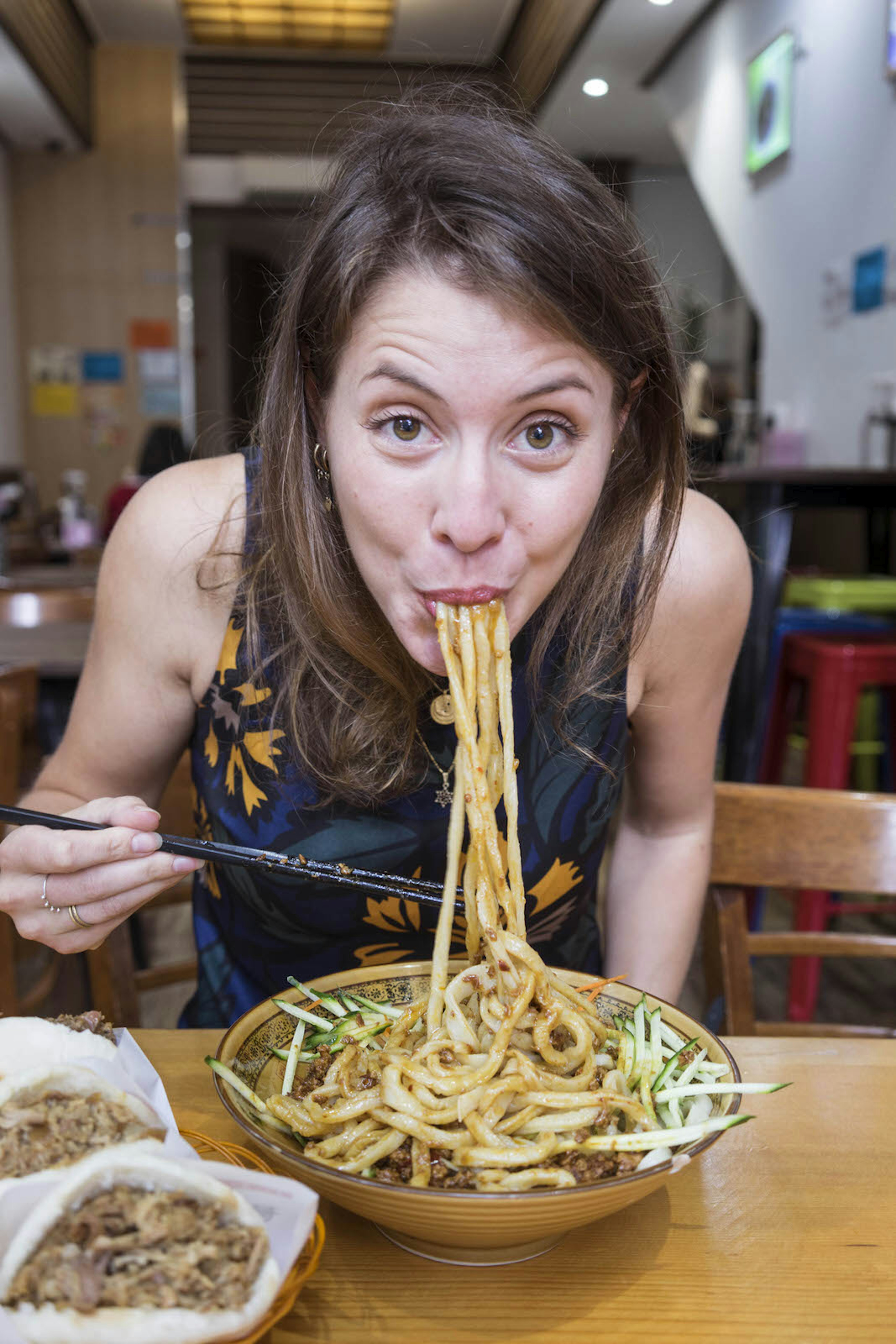 Sofia Levin biting into a mouthful of saucy noodles, looking into the camera with chopsticks in her hand. There are two other side dishes on the table and the restaurant is blurred in the background.