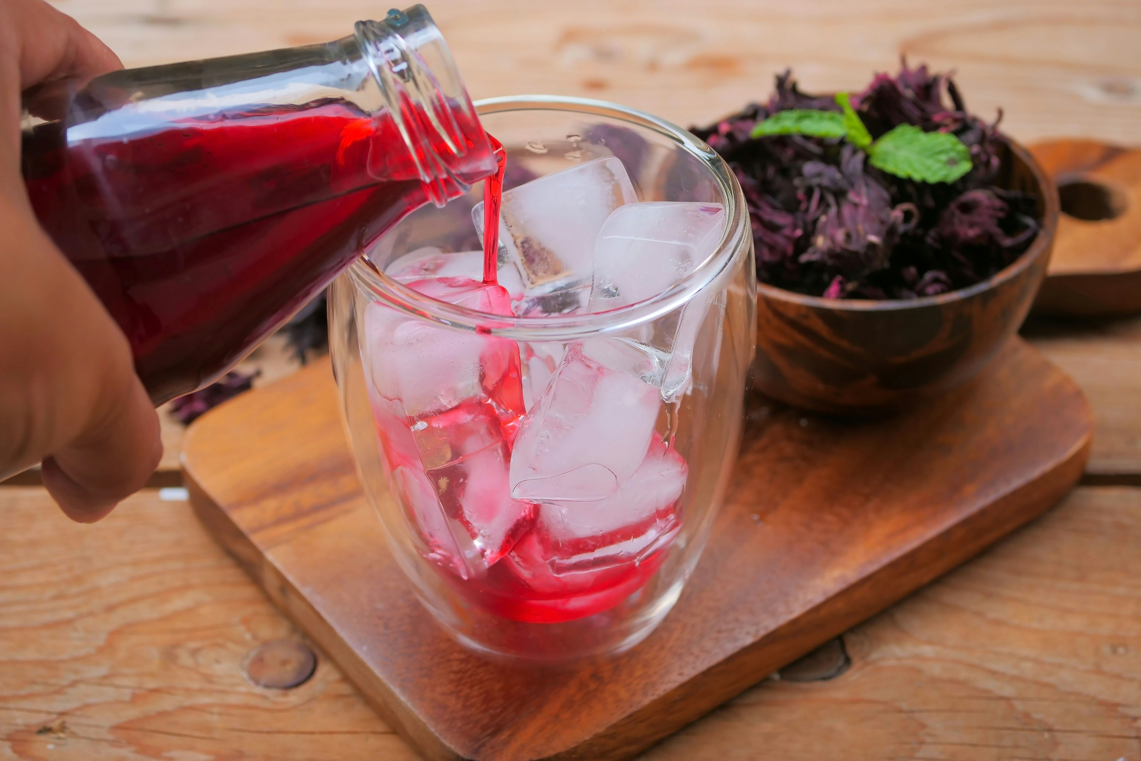 A person pours crimson-colored sorrel into a glass filled with ice. In the background is a bowl of dried sorrel topped with a green herb.