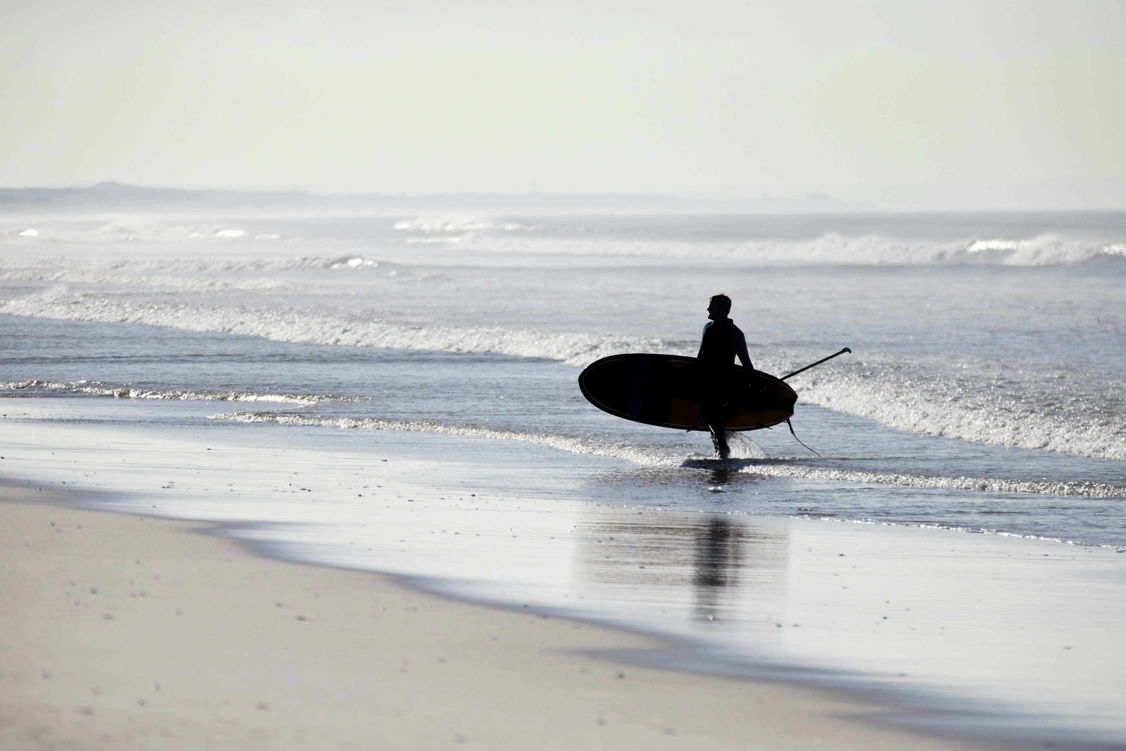 Paddleboarder walking out of ocean carrying paddleboard; places to go stand up paddle boarding