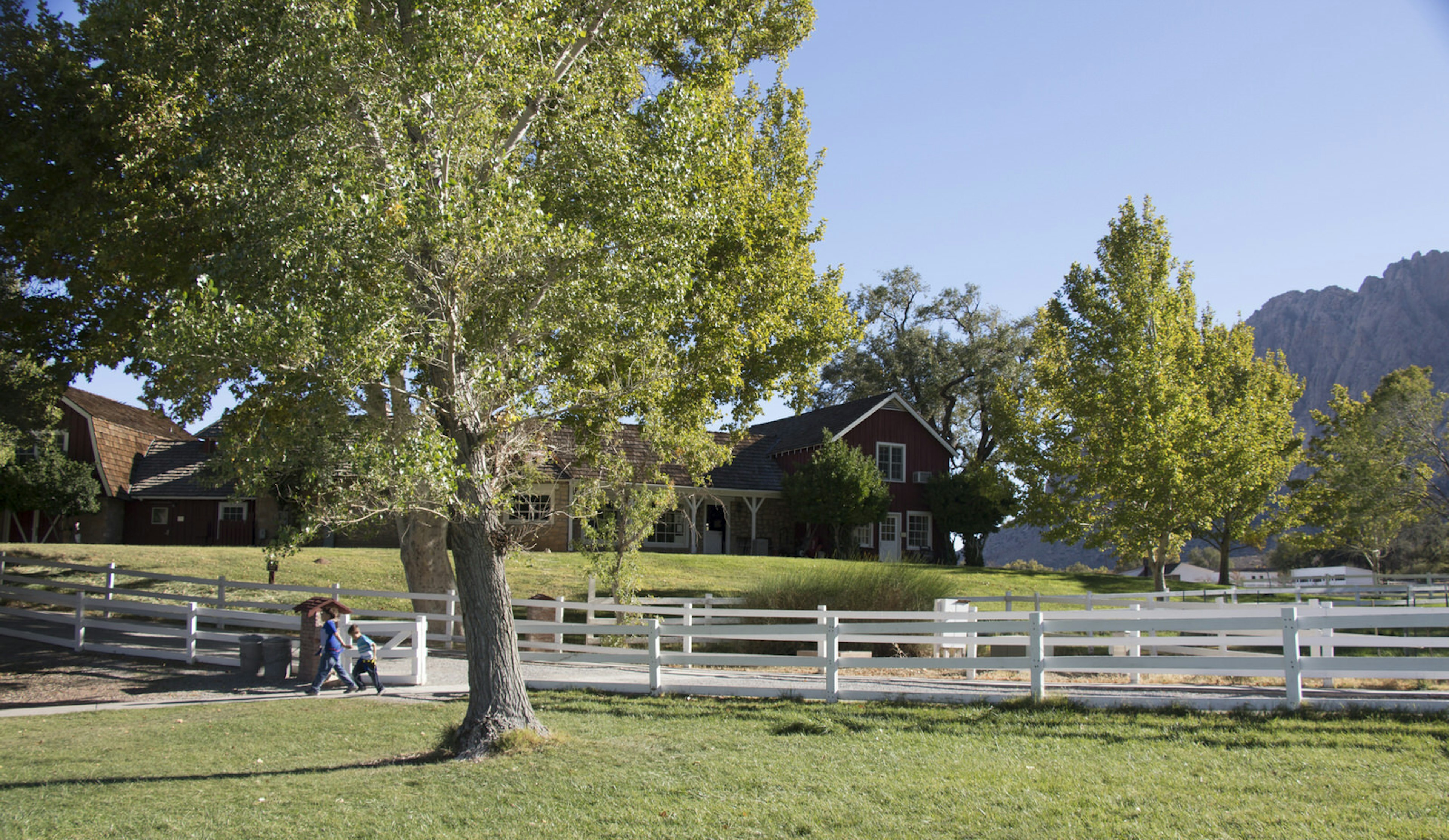 A lovely red ranch house stands at the top of a grassy green hill, with white picket fences outlining a dirt drive leading to it. People walk along the drive, looking at the rocky outcrops of mountains in the background © Greg Thilmont / Lonely Planet