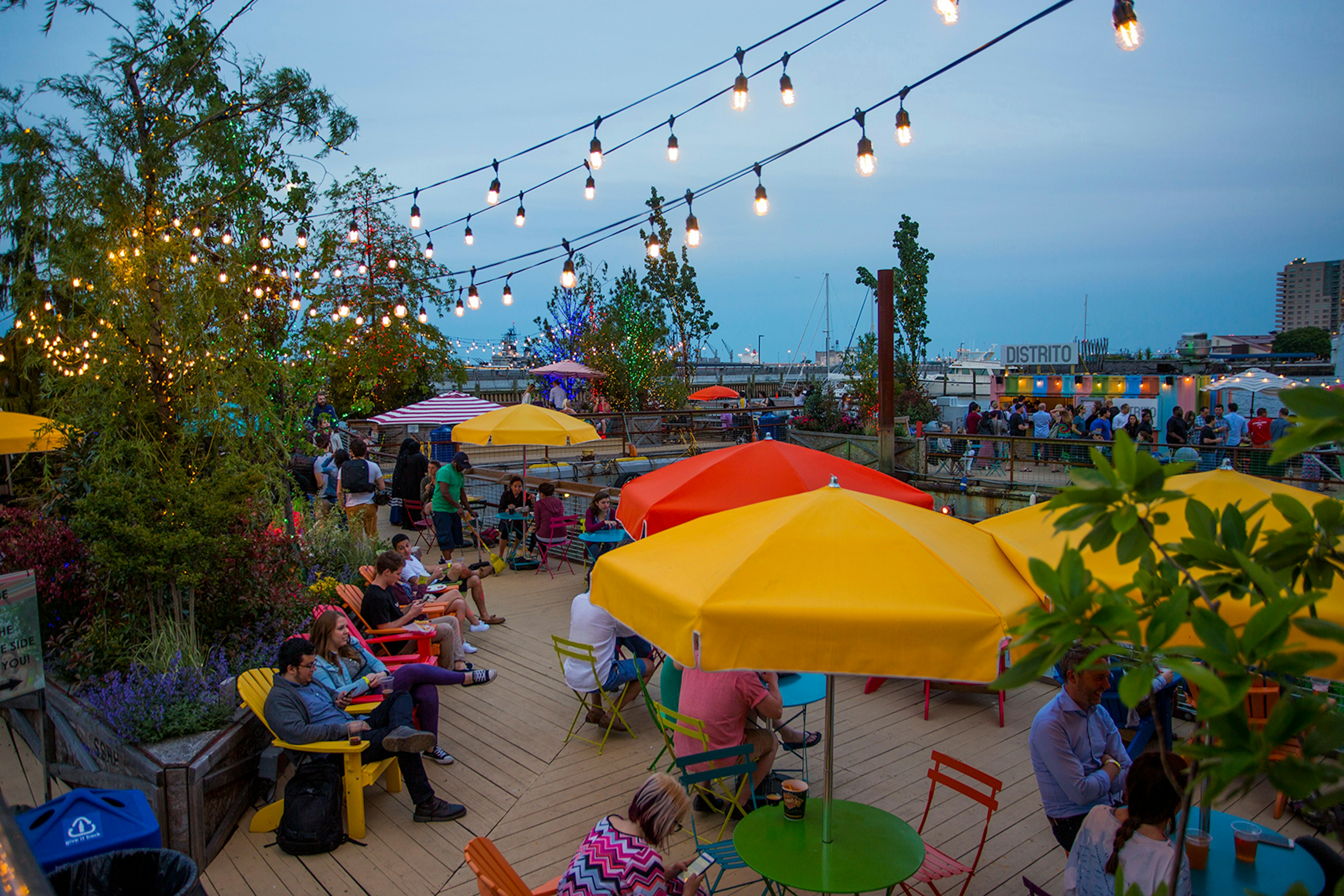 Overhead view of a city park, with tables shaded by bright yellow and red umbrellas and Edison bulb fairy lights overhead