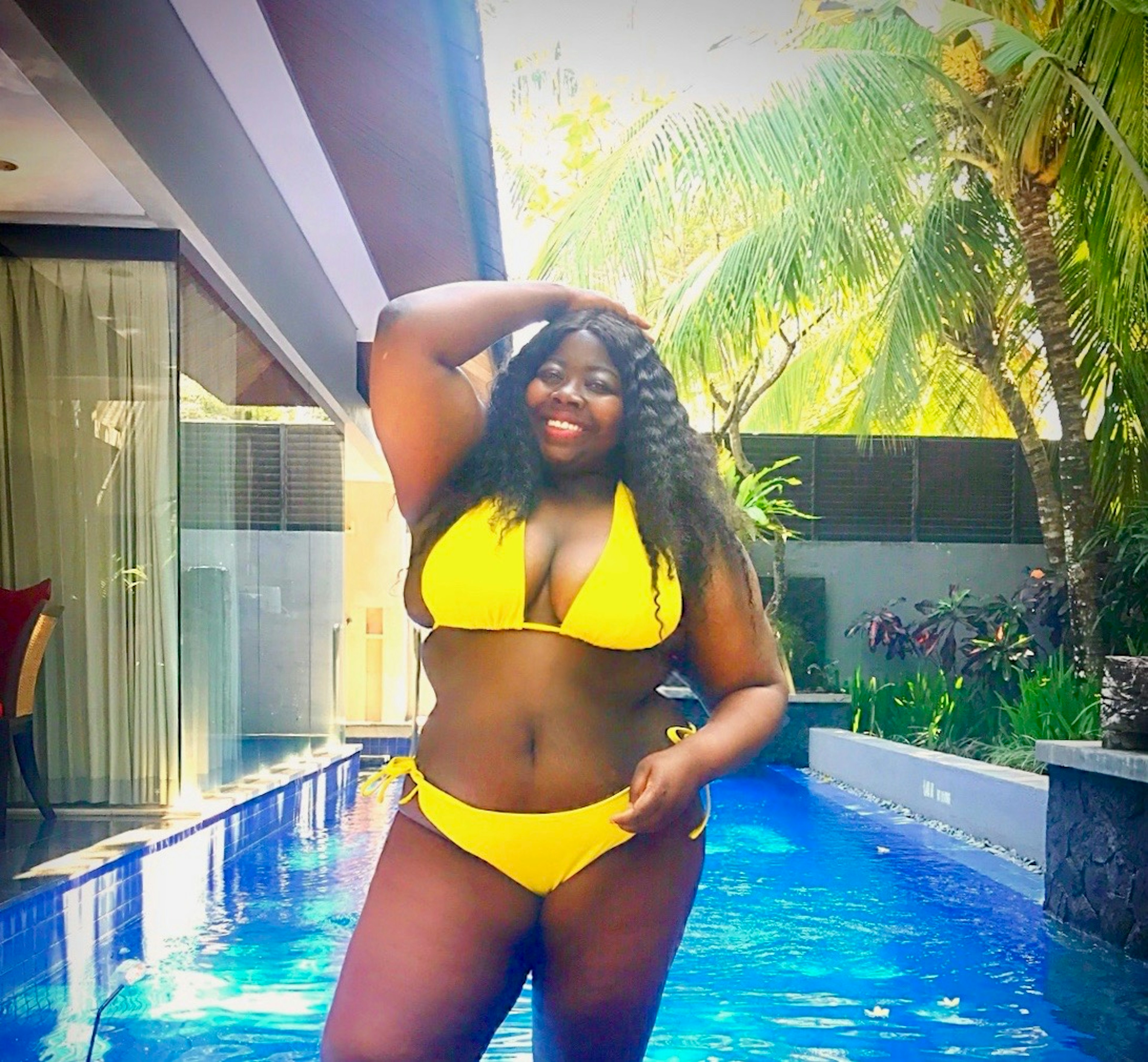 A black plus size woman poses in front of a private pool in a yellow bikini. The sun is streaming through the palm trees, she's smiling and looks really relaxed and happy. A black girl travelling in Bali tip: plus-size woman attract a lot of attention.