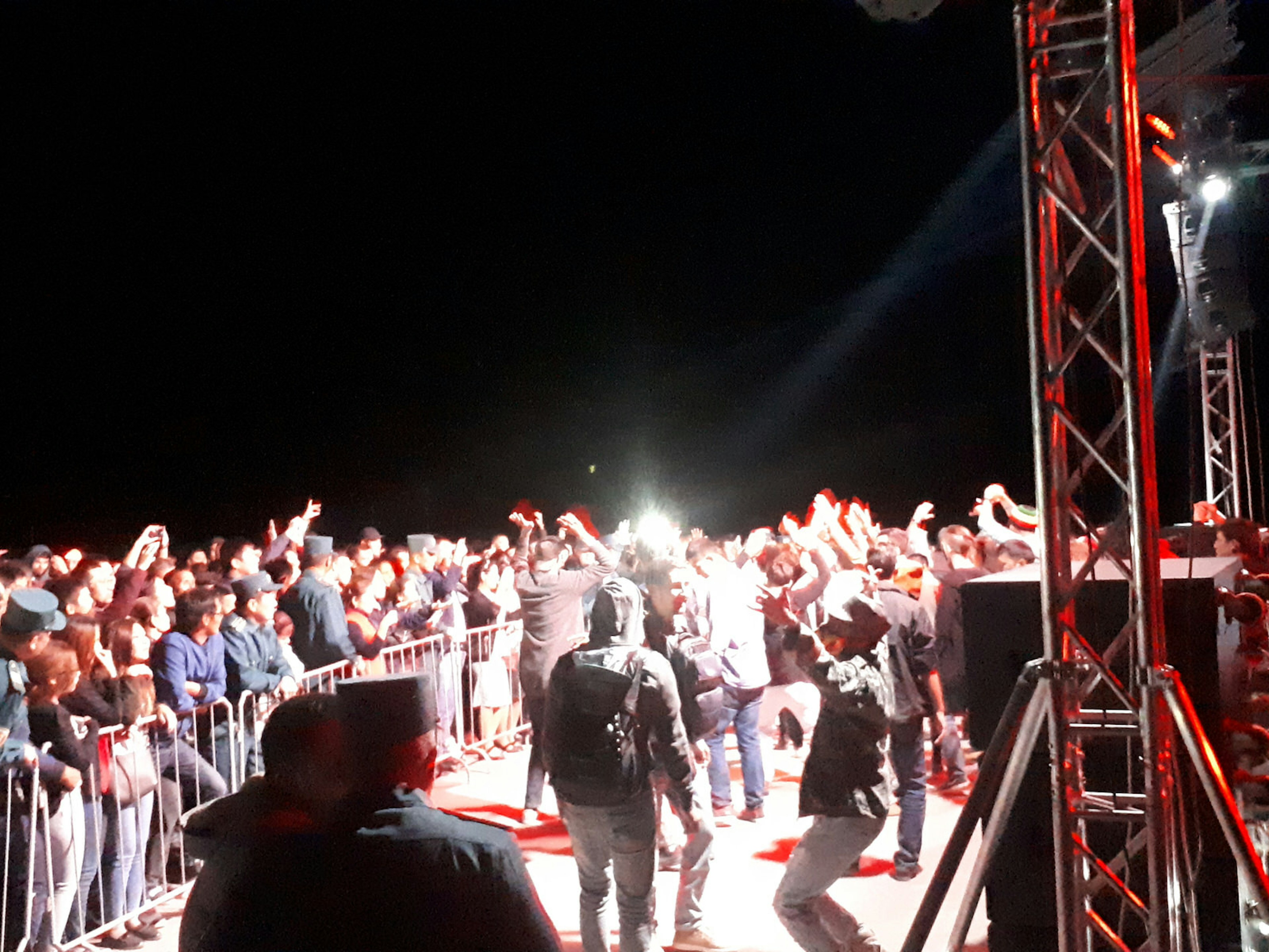 A large audience with hands raised in a night-time scene next to an illuminated stage