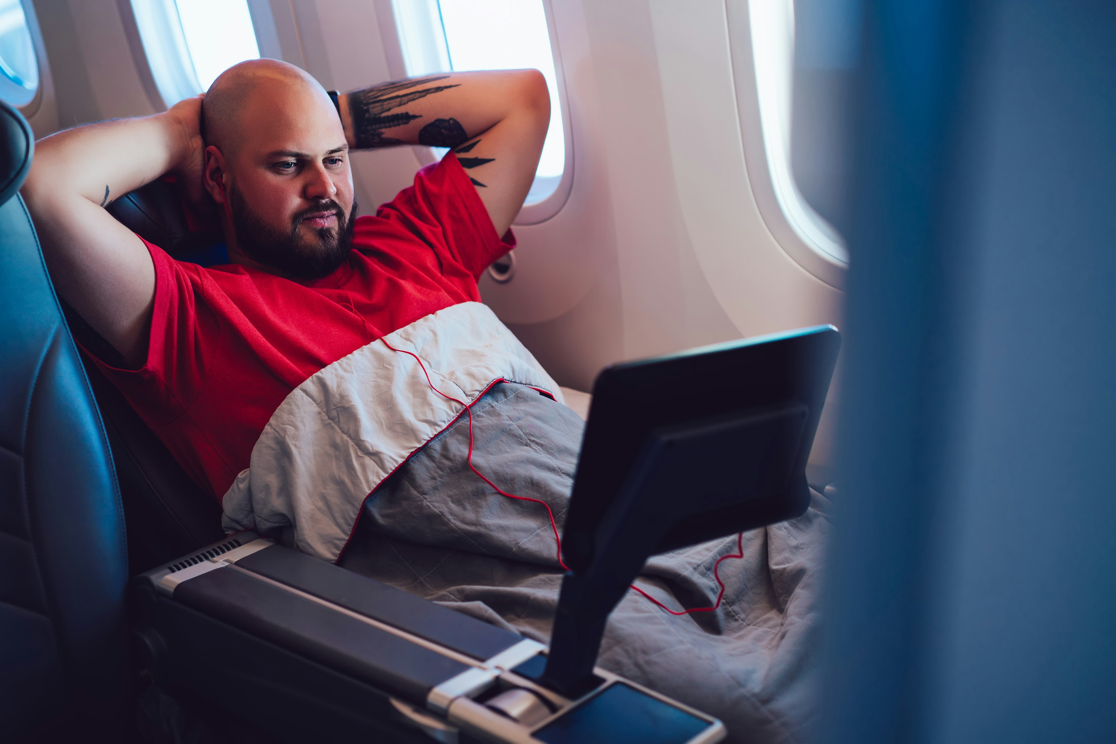 A man reclines in an airplane seat with his hands folded behind his head as he watches a small screen. He's got a blanket pulled up to the middle of his chest and is next to the window.